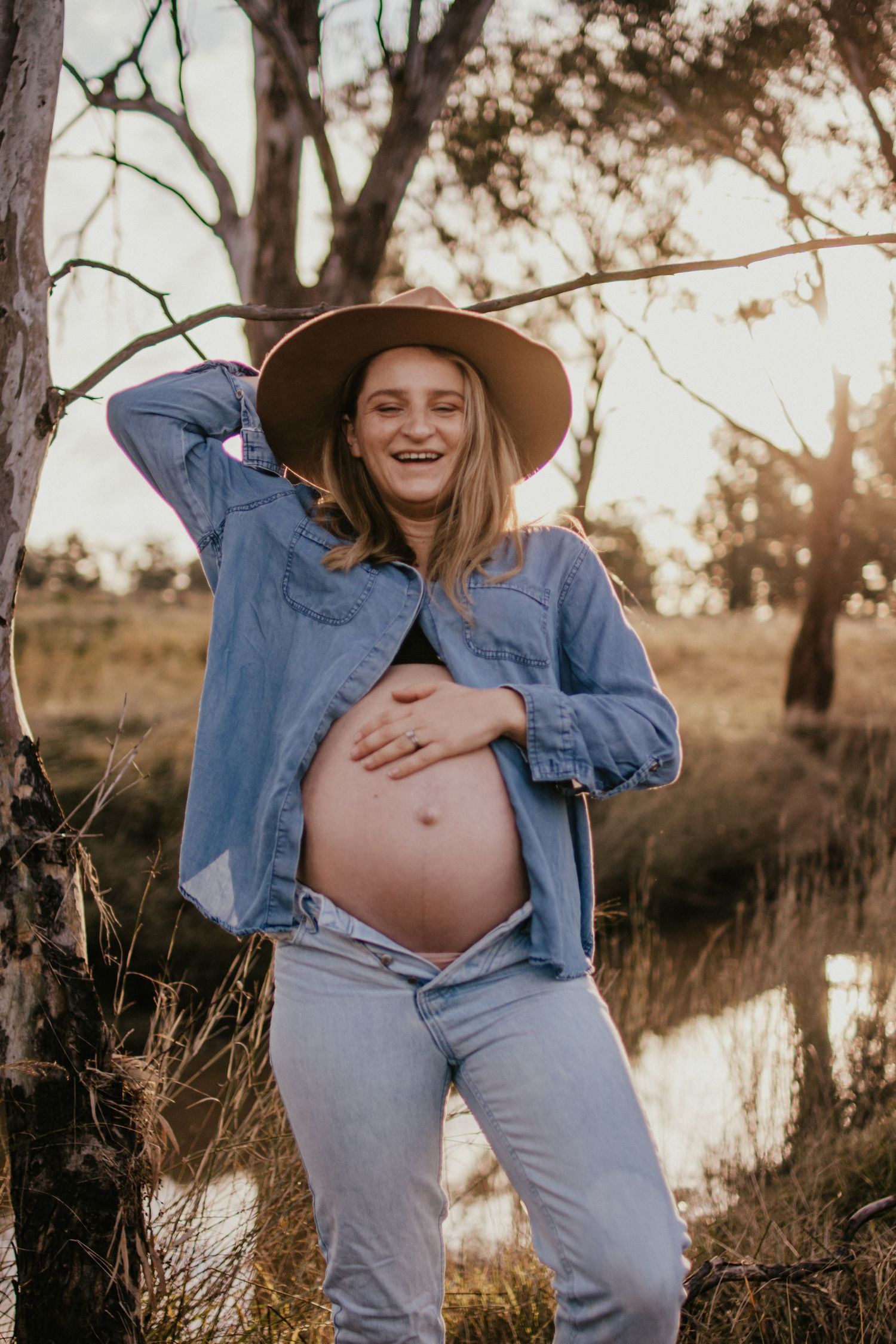 Pregnant lady in a denim shirt with beautiful baby bump showing. Standing in a paddock with a creek in the background.