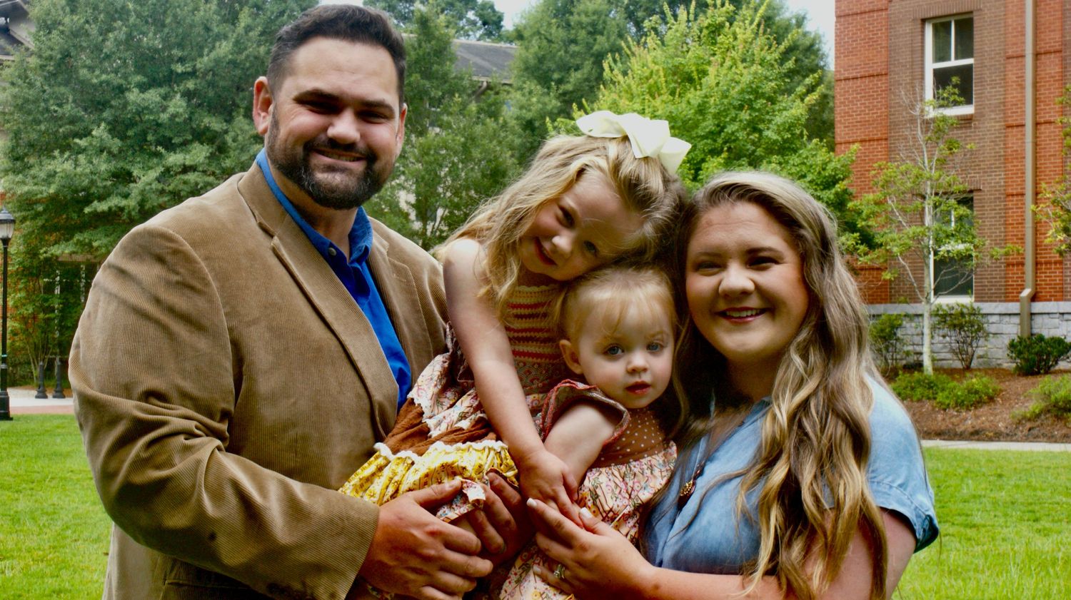 Happy family of three smiling together on grassy lawn with brick building in background.
