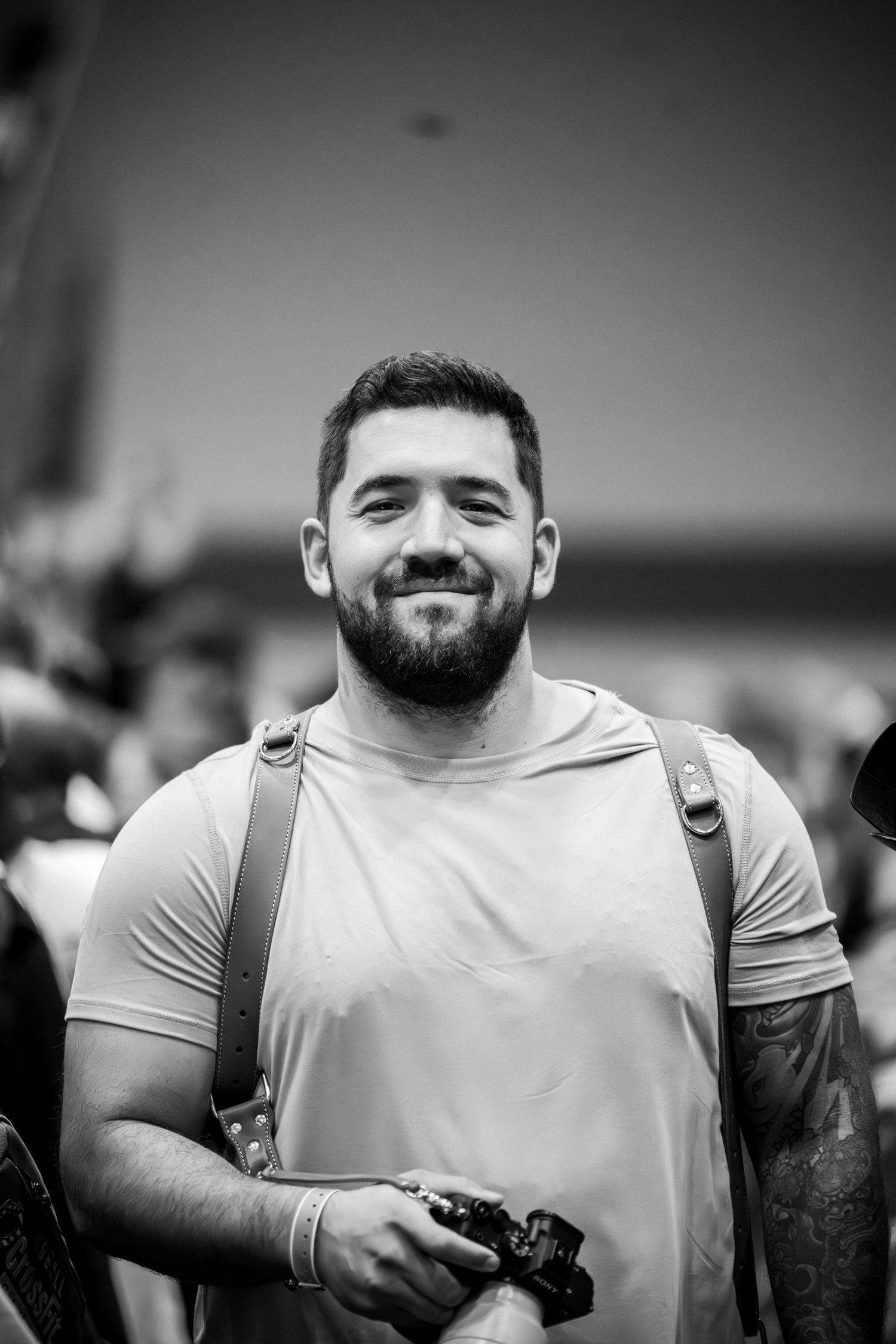 Bearded athlete in light gray t-shirt holding camera at indoor sports event.