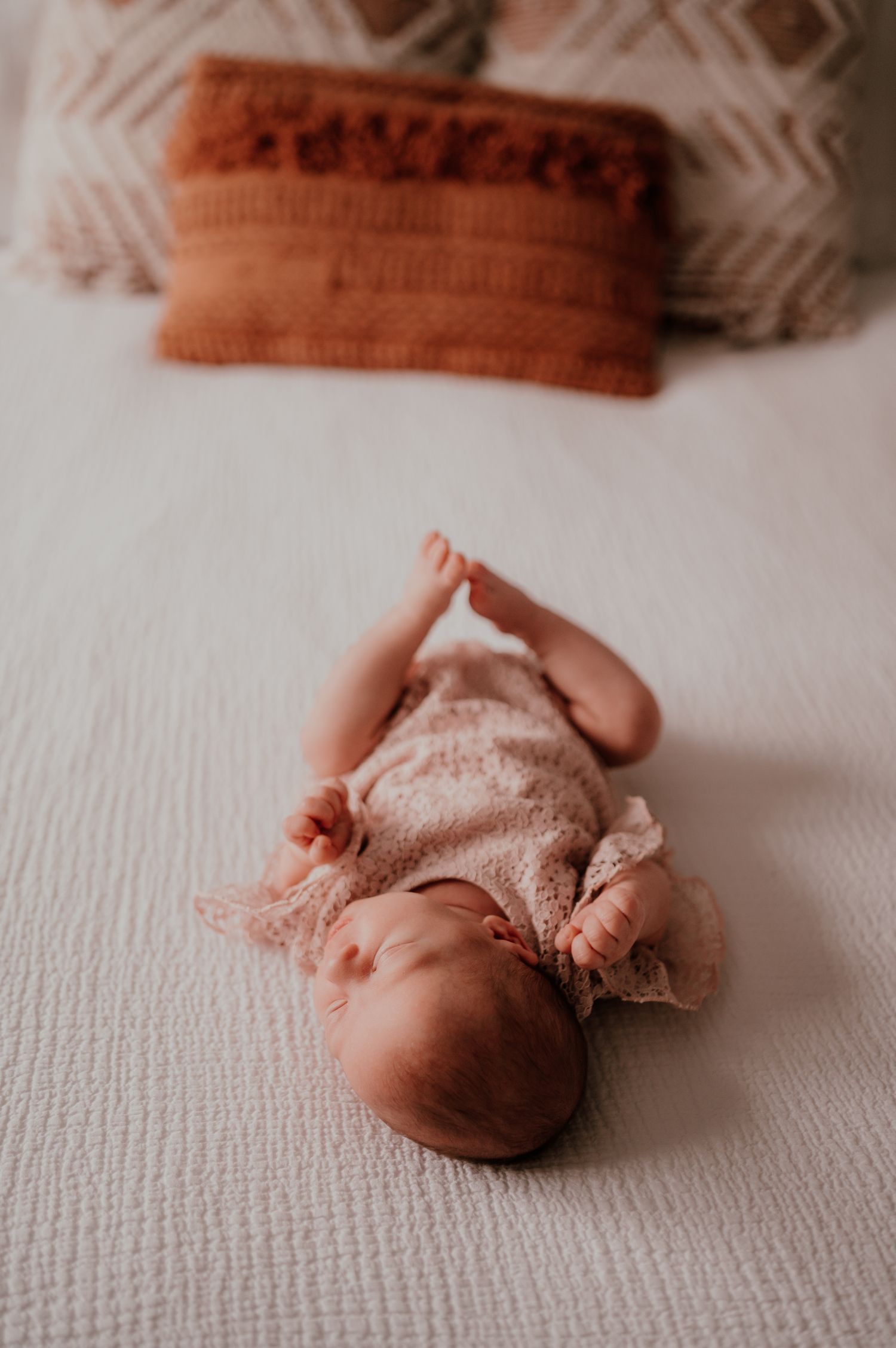 A sleeping baby in lace outfit lying on white bedding with orange pillow above.