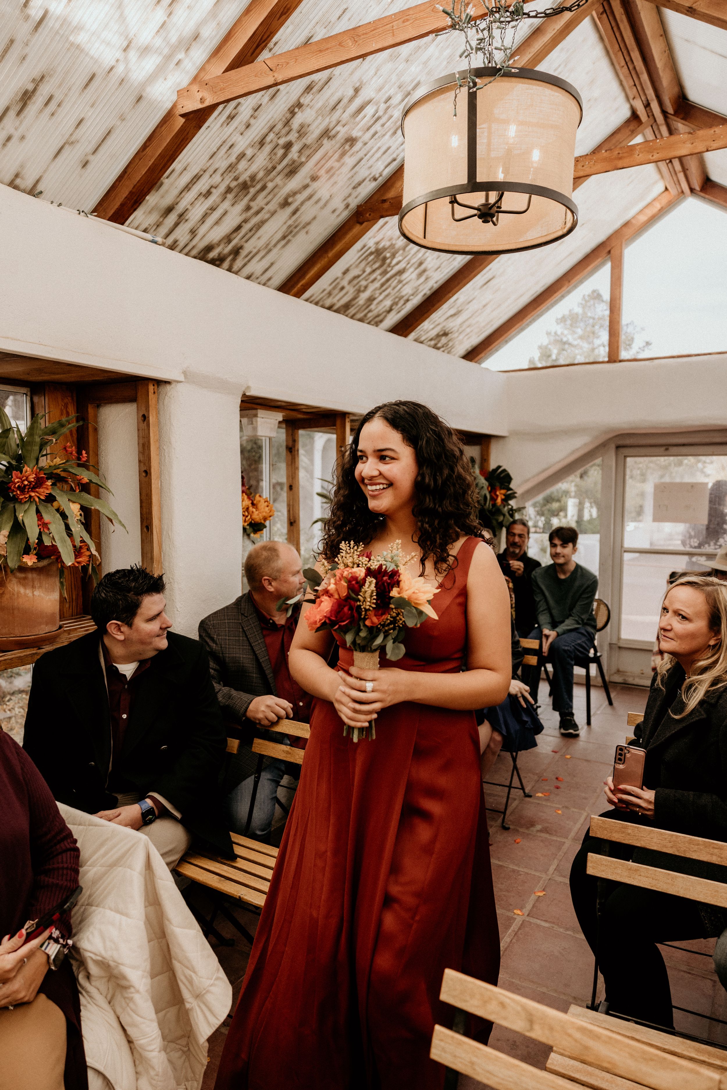 Smiling bridesmaid in rust red gown walks down aisle holding autumn floral bouquet.