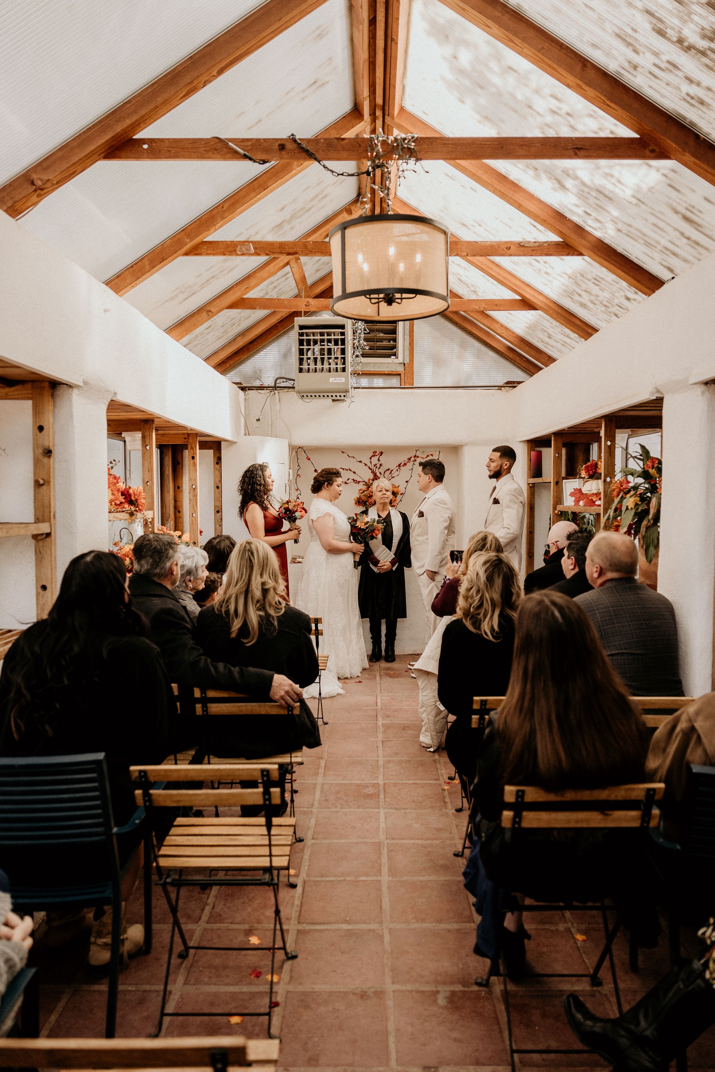 Couple exchanges vows in warmly lit barn wedding venue with exposed beams and fall floral decor.