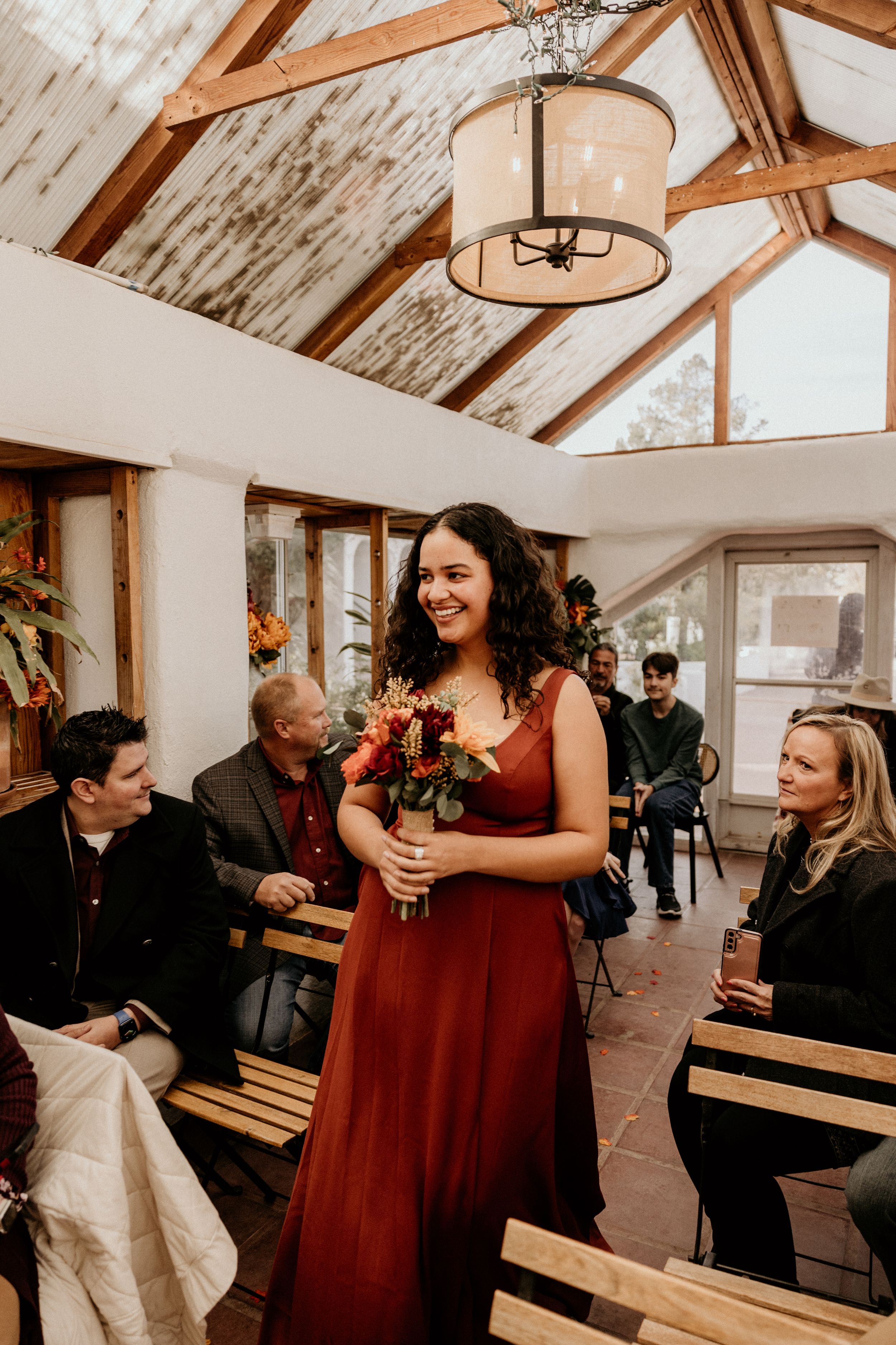 Bridesmaid in burgundy dress smiles while walking down aisle with fall bouquet in barn venue.