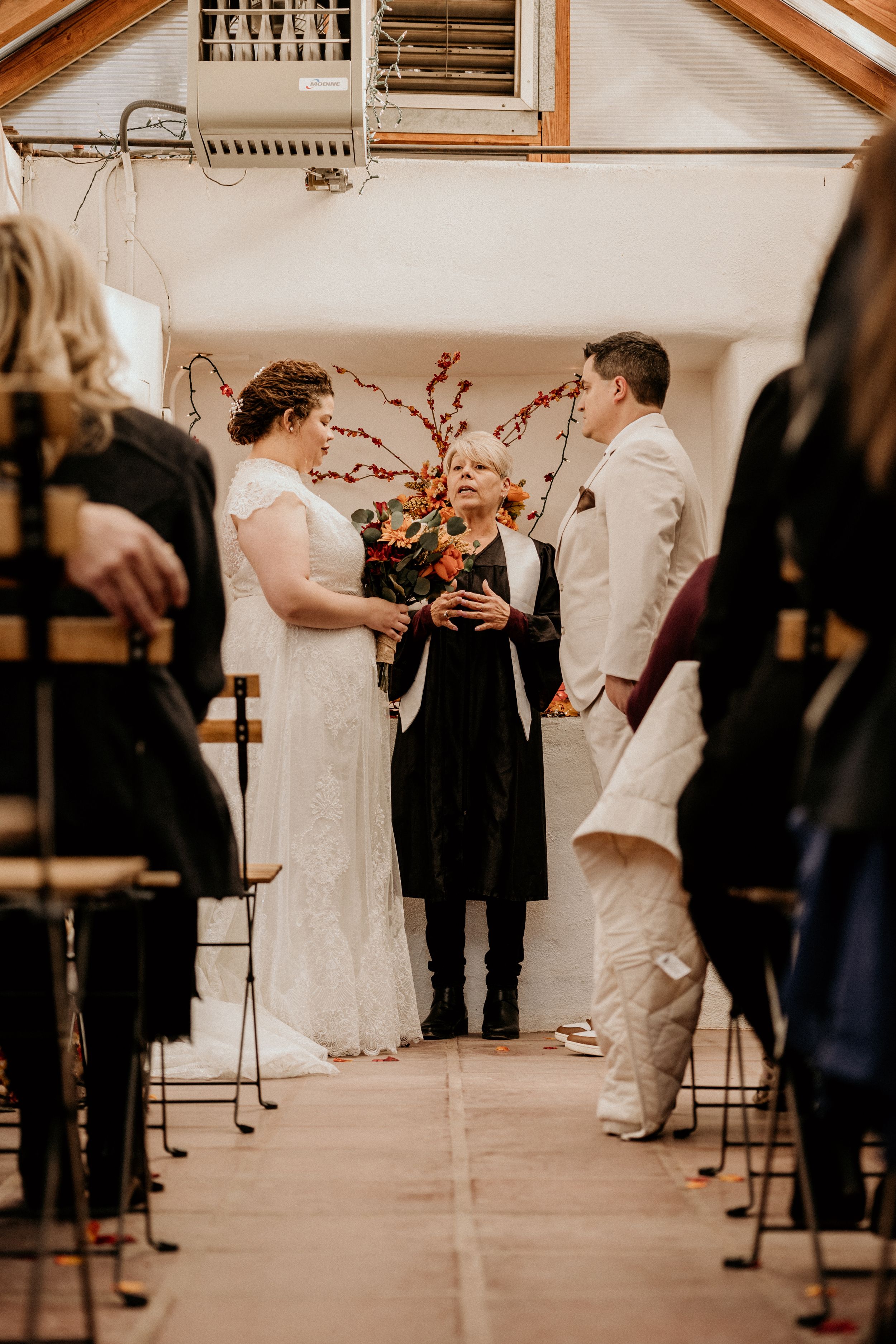 Officiant speaks to bride in lace gown and groom in white suit during intimate barn wedding ceremony.
