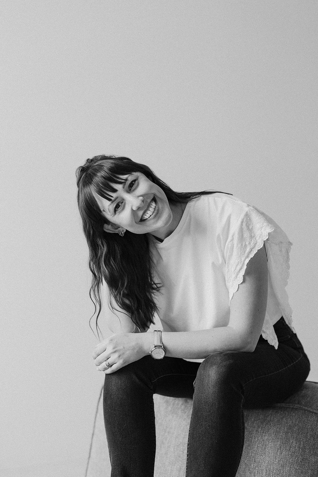 Casual portrait of someone in a white top sitting and smiling against a grey background.