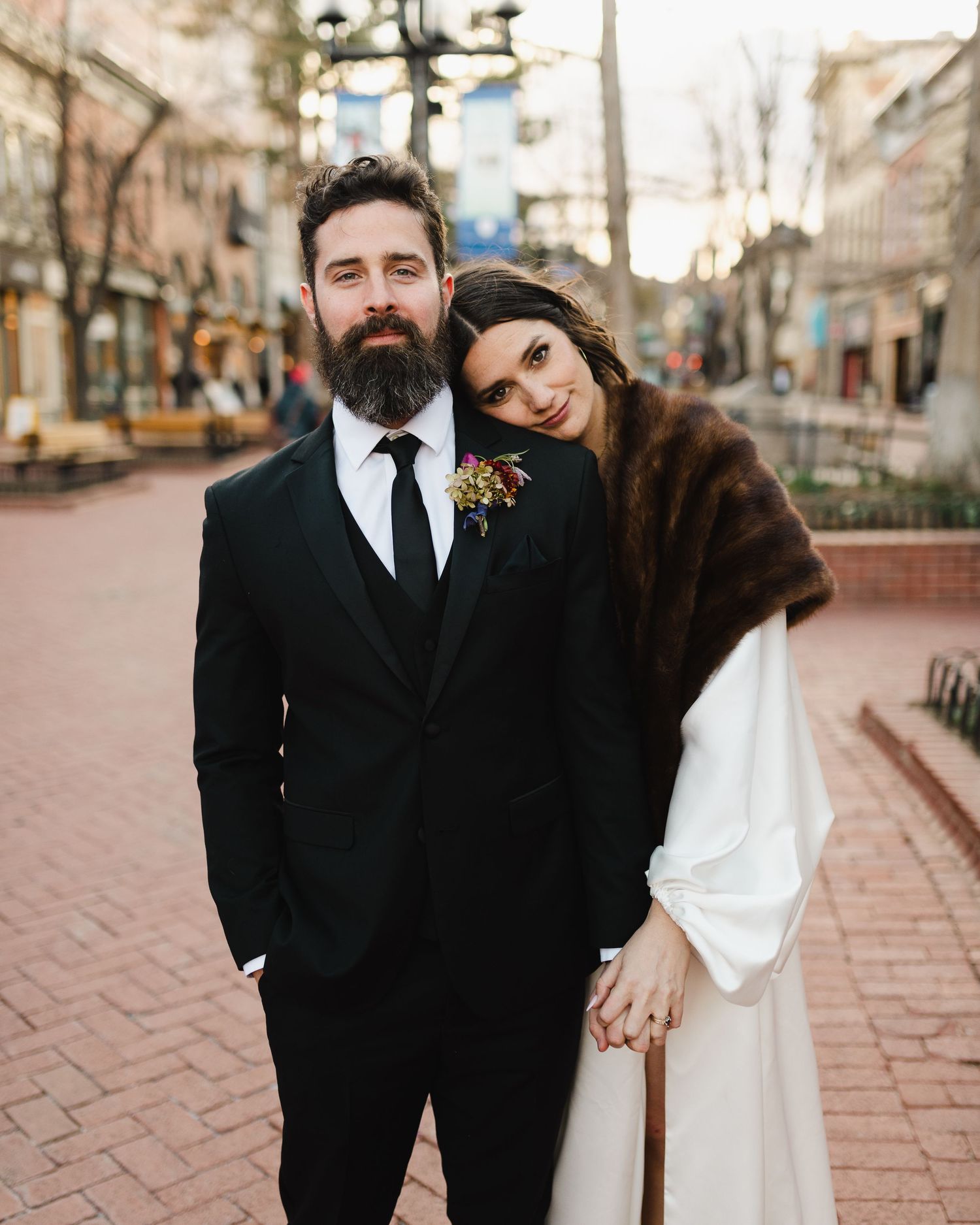 A stylish wedding couple poses on a brick pathway, with the bride wearing a fur stole and the groom in a black suit.