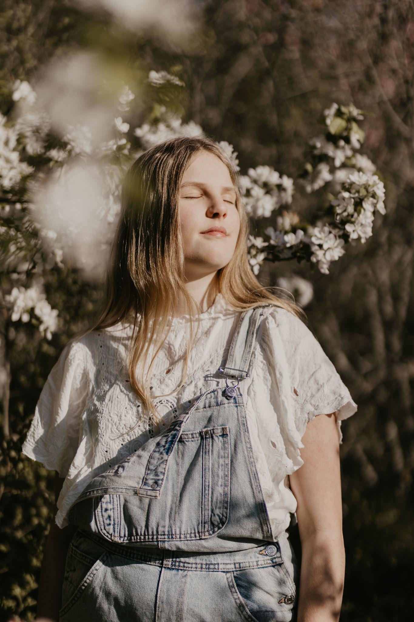 Person in denim overalls and white shirt standing among spring blossoms with eyes closed.
