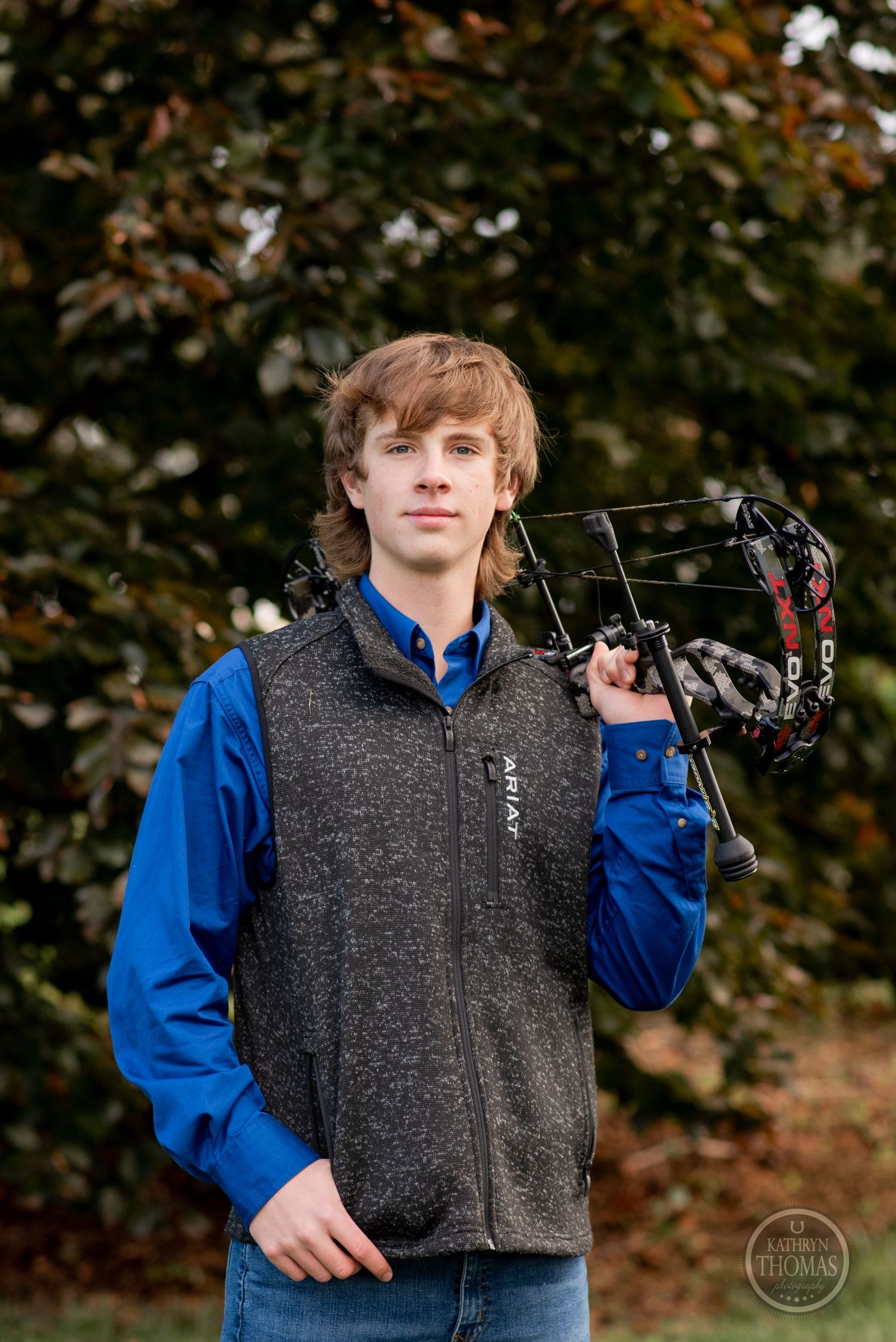 An archer poses with a compound bow against autumn foliage wearing blue and gray.