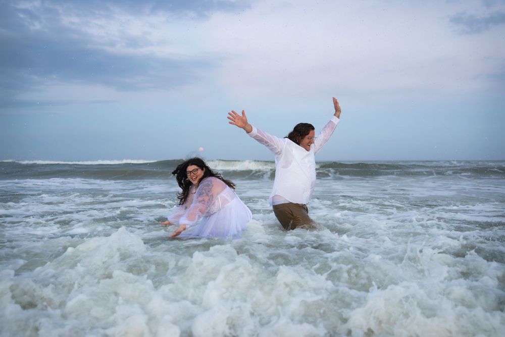 A couple splashes joyfully in ocean waves while wearing wedding attire.
