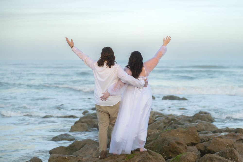 Two people raise their arms in celebration while standing on coastal rocks at sunset.