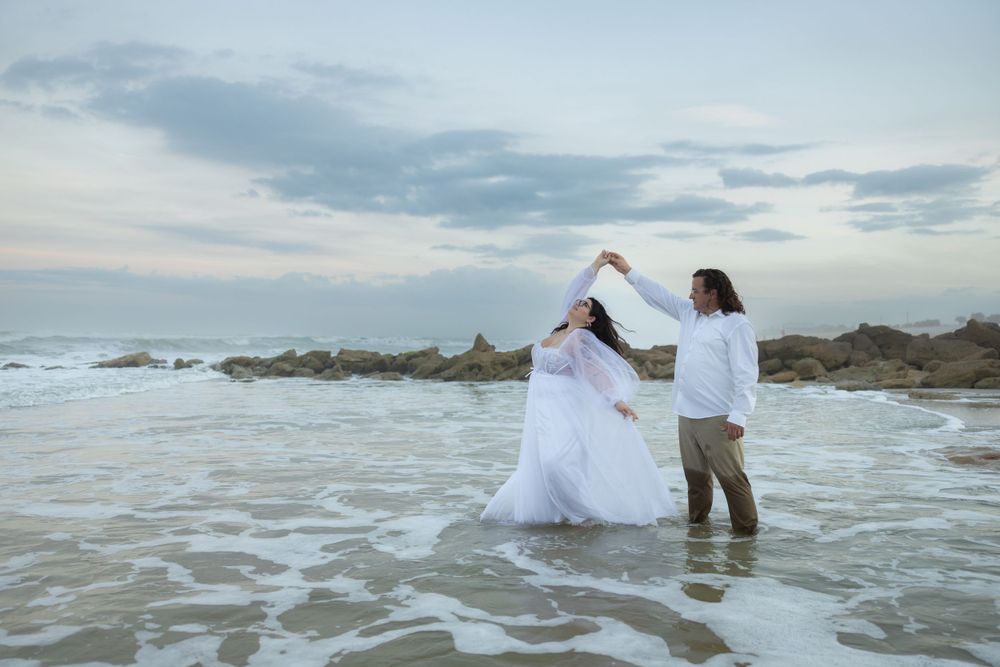 A couple dancing together in white attire on a rocky beach at dusk.