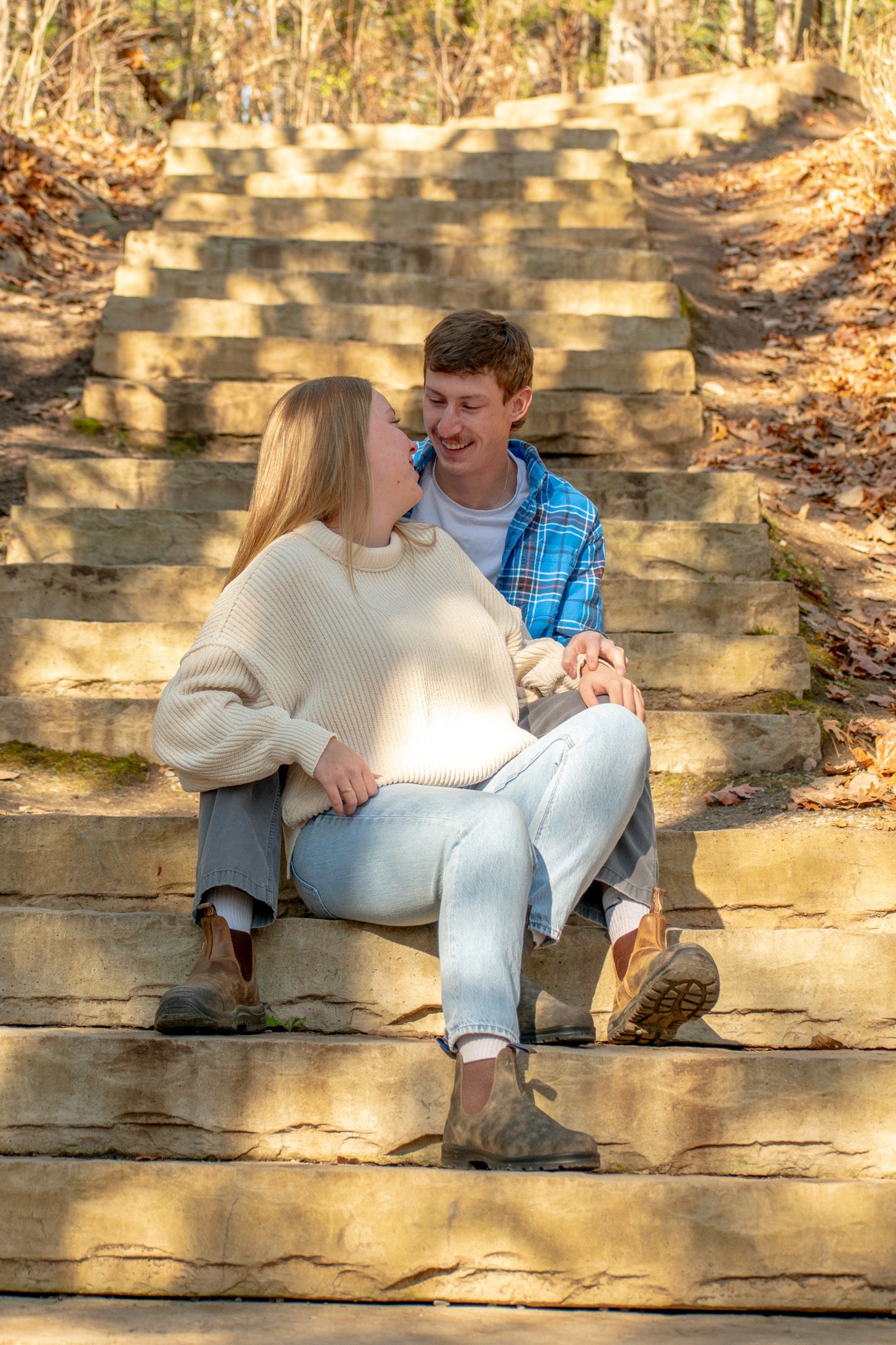 A couple sits together on stone steps surrounded by fall foliage on a sunny autumn day.
