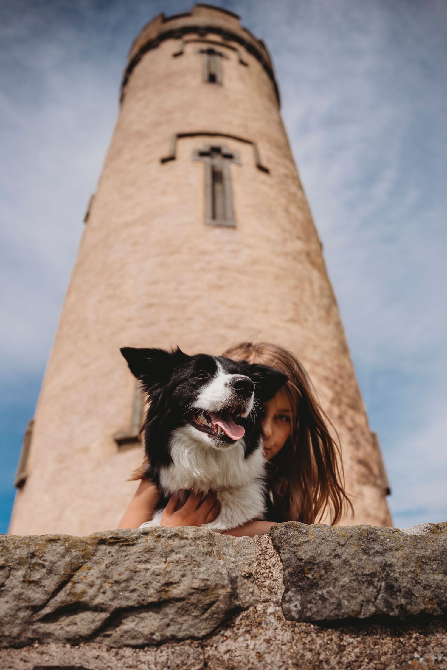 A border collie poses in front of a tall stone lighthouse against a cloudy blue sky.