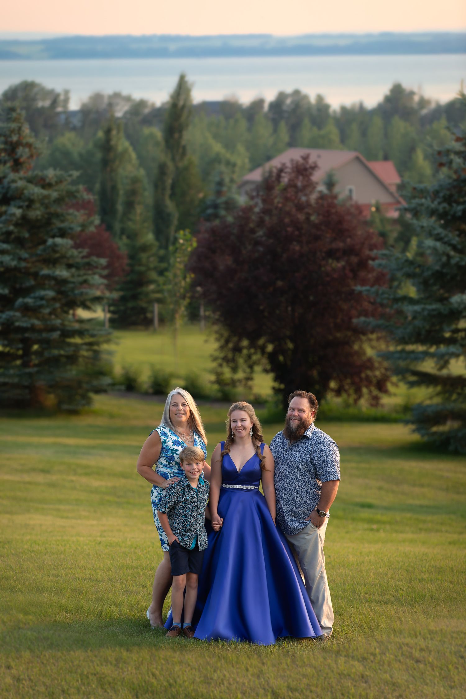A group stands on a grassy hillside at sunset with evergreen trees and a lake visible in the background.