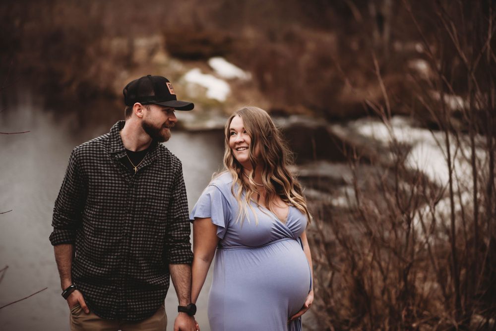 A couple walks along a waterfront path during maternity photoshoot.