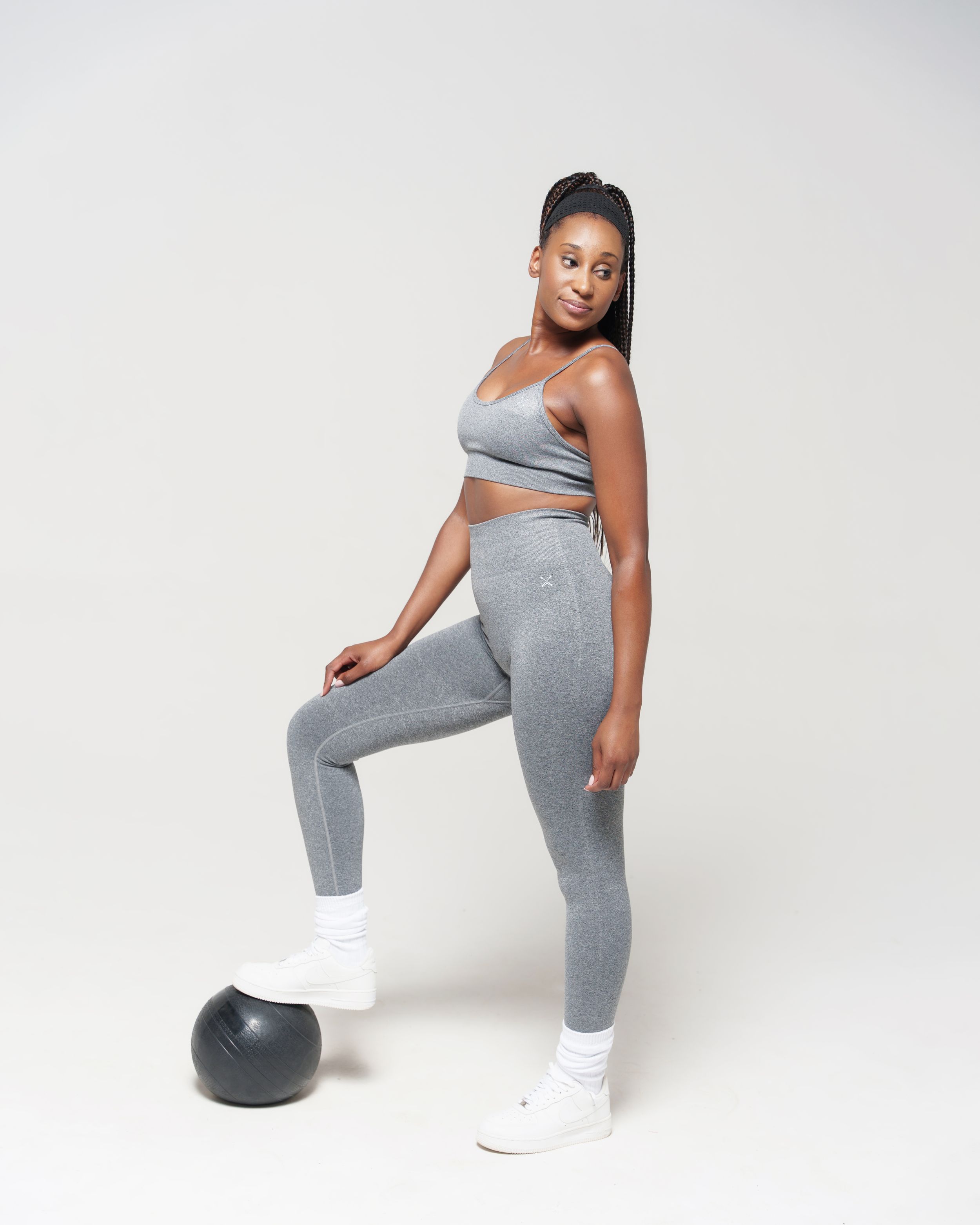 Athletic model in gray workout set poses with foot on exercise ball against white background.