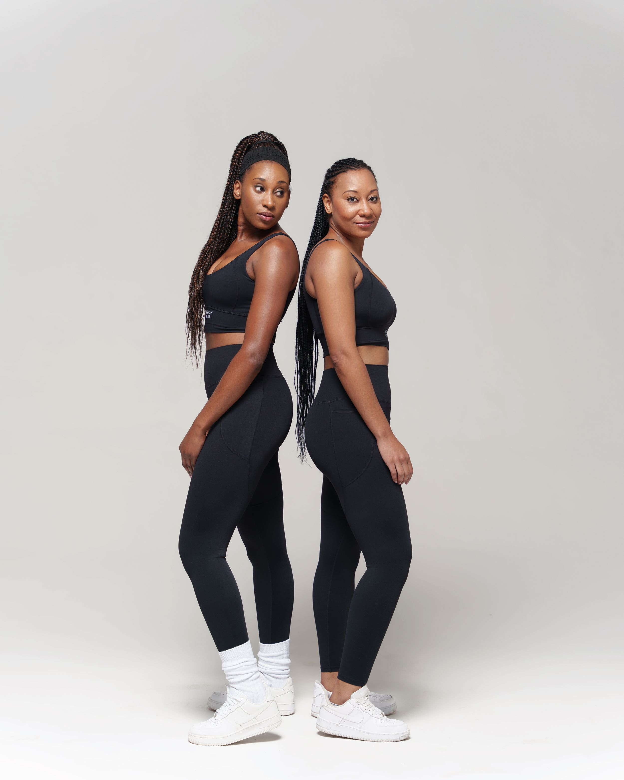 Two models pose back to back wearing matching black athletic sets and white sneakers against a light gray backdrop.