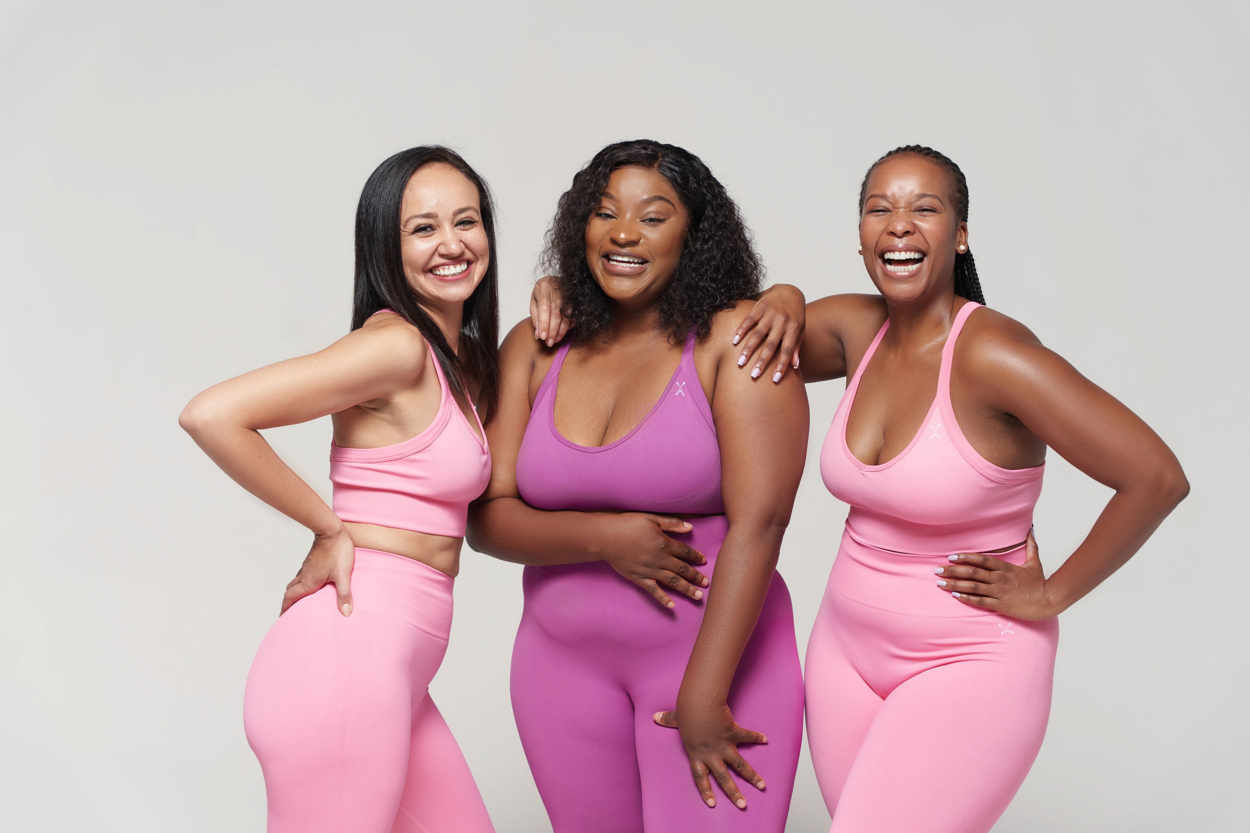 Three women in pink and purple workout sets smile and pose together against a white background.