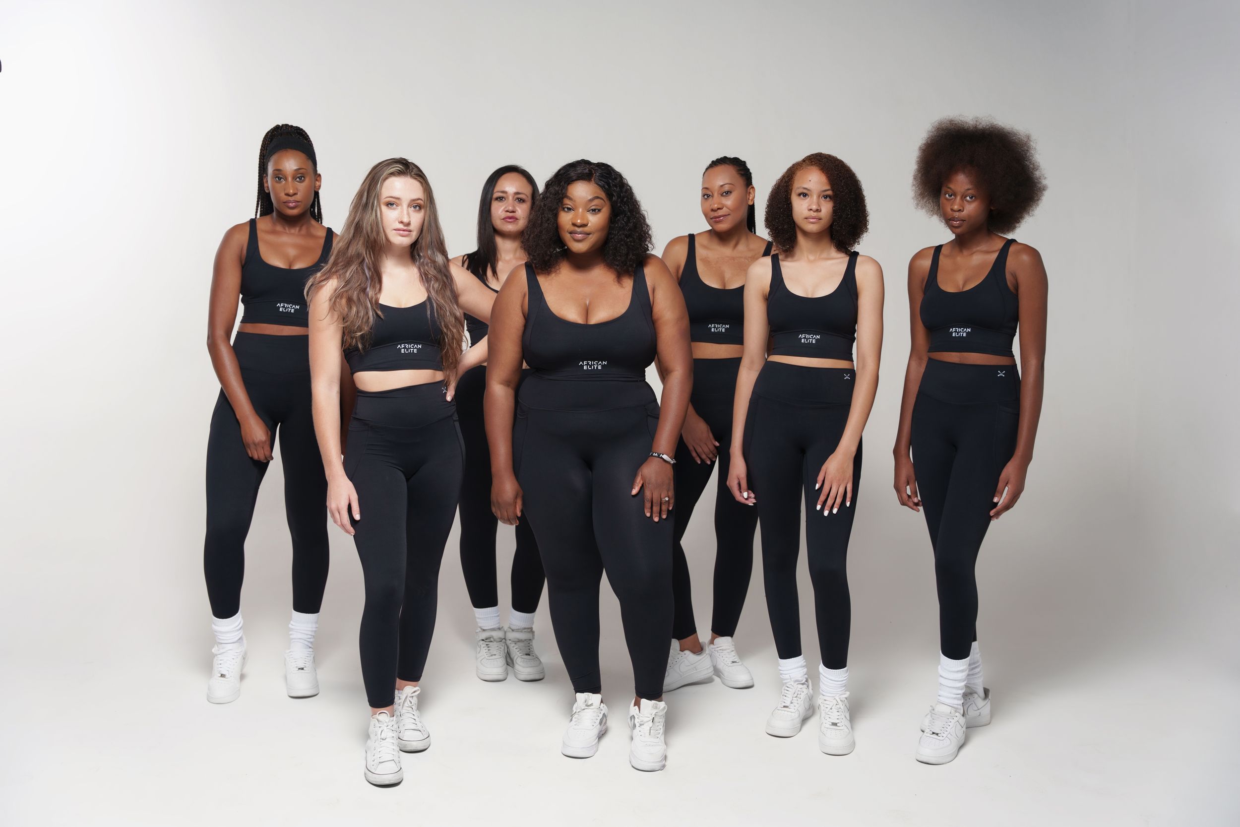 Group of diverse models standing together wearing matching black athletic wear and white sneakers.