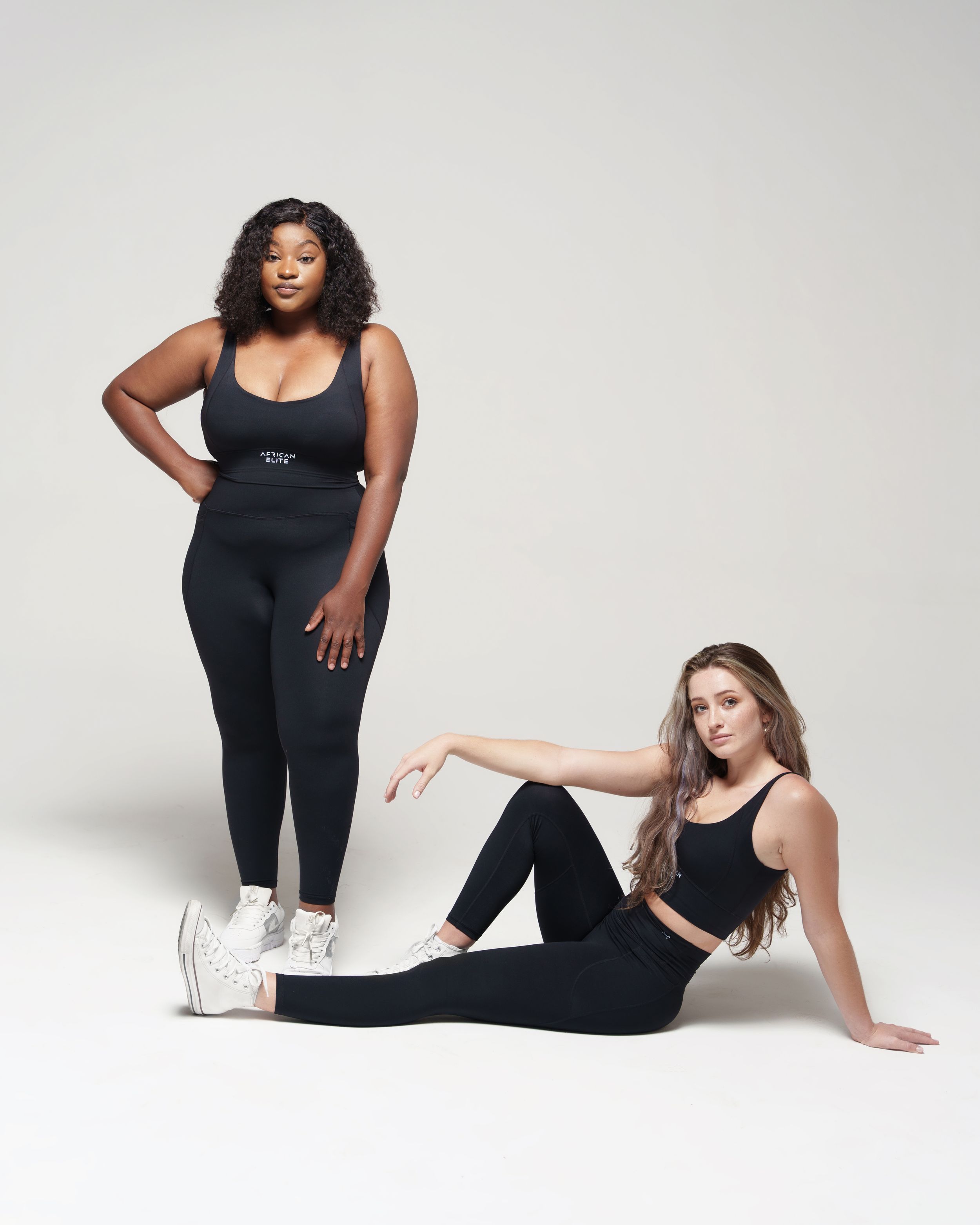 Two models in black activewear pose together, one standing and one seated on the floor.
