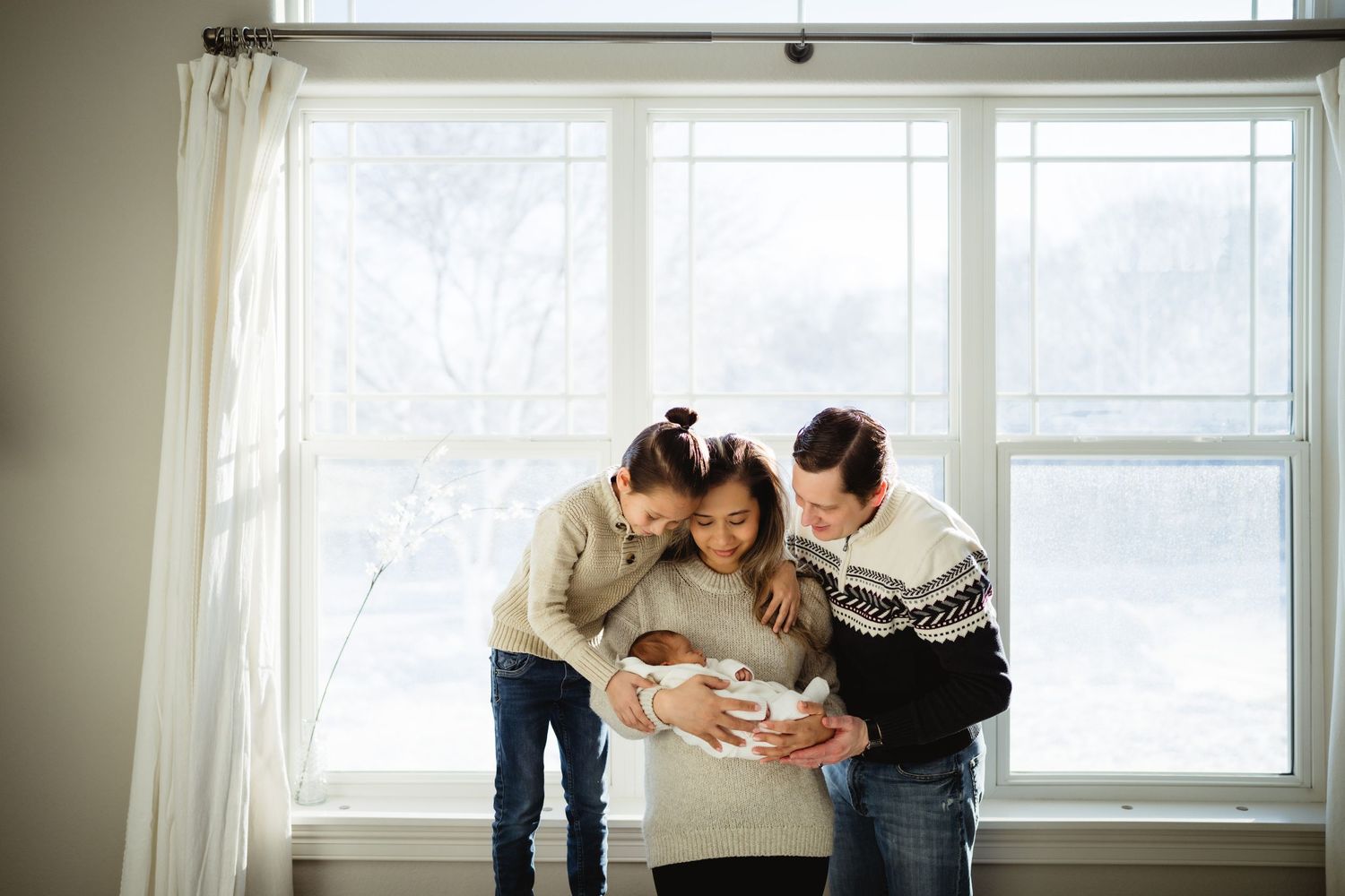 A family gathers by a bright window to admire their newborn baby.