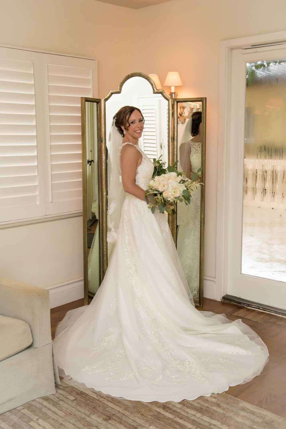 Bride shows off stunning lace wedding dress with cathedral train in sunlit room.