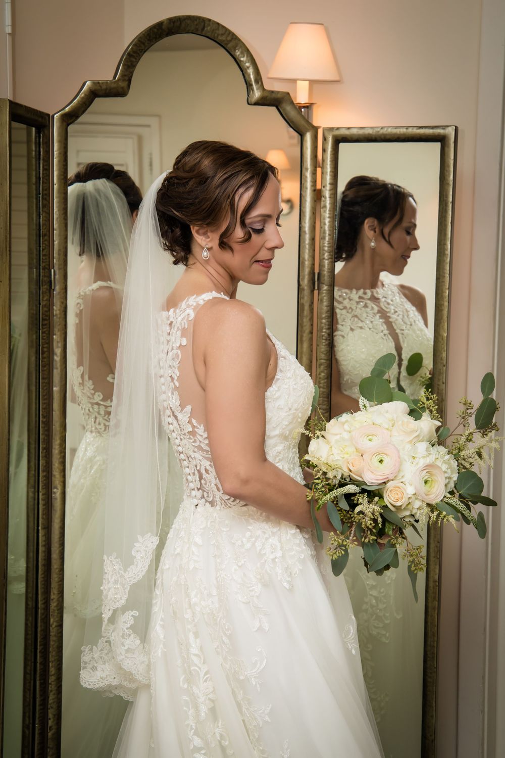 Bride holding white bouquet poses in front of mirror wearing detailed lace wedding gown.