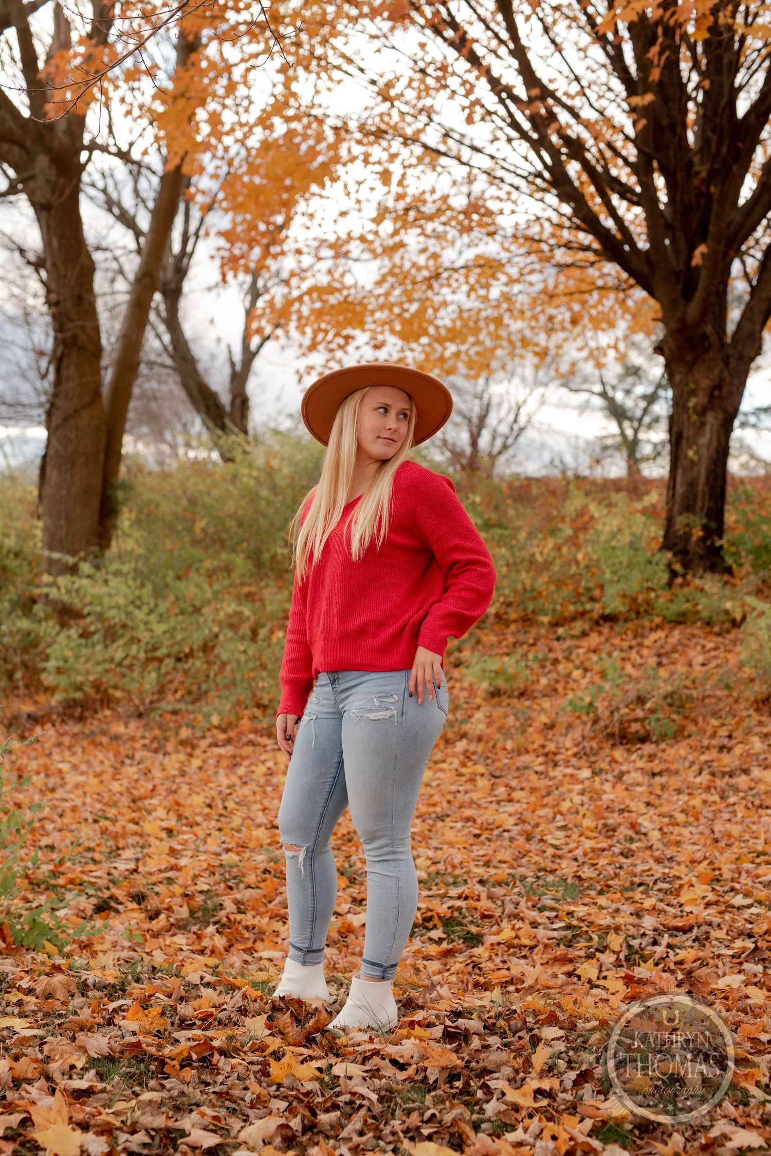 Fall fashion portrait with red sweater, wide brim hat and light denim against autumn trees.