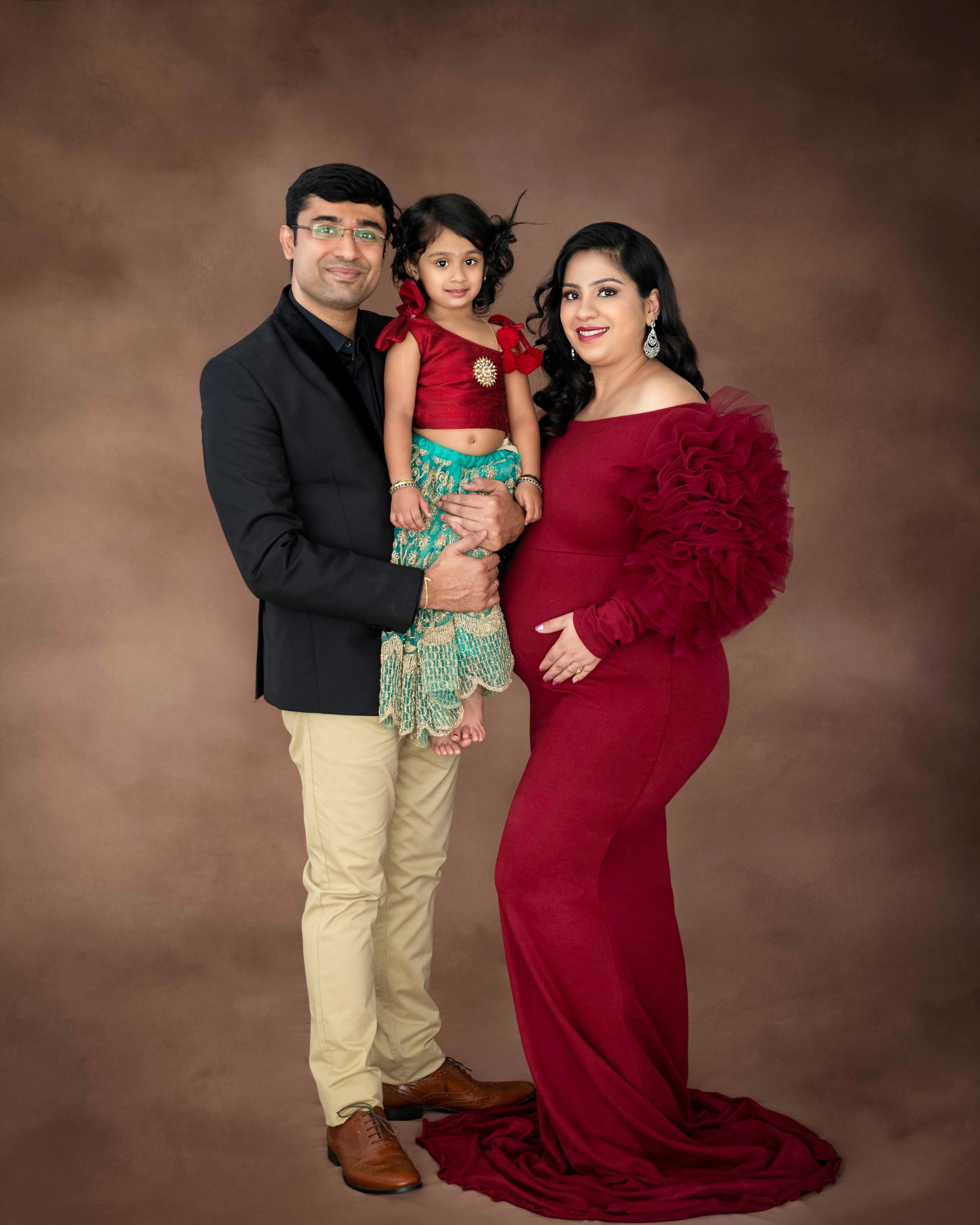 Studio family portrait with parents and child wearing coordinated red and beige outfits against brown backdrop.