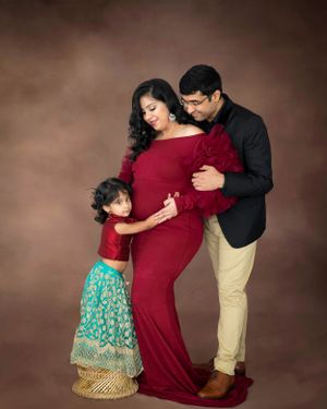Family maternity photo with a young child in traditional dress embracing expecting mother in red gown.