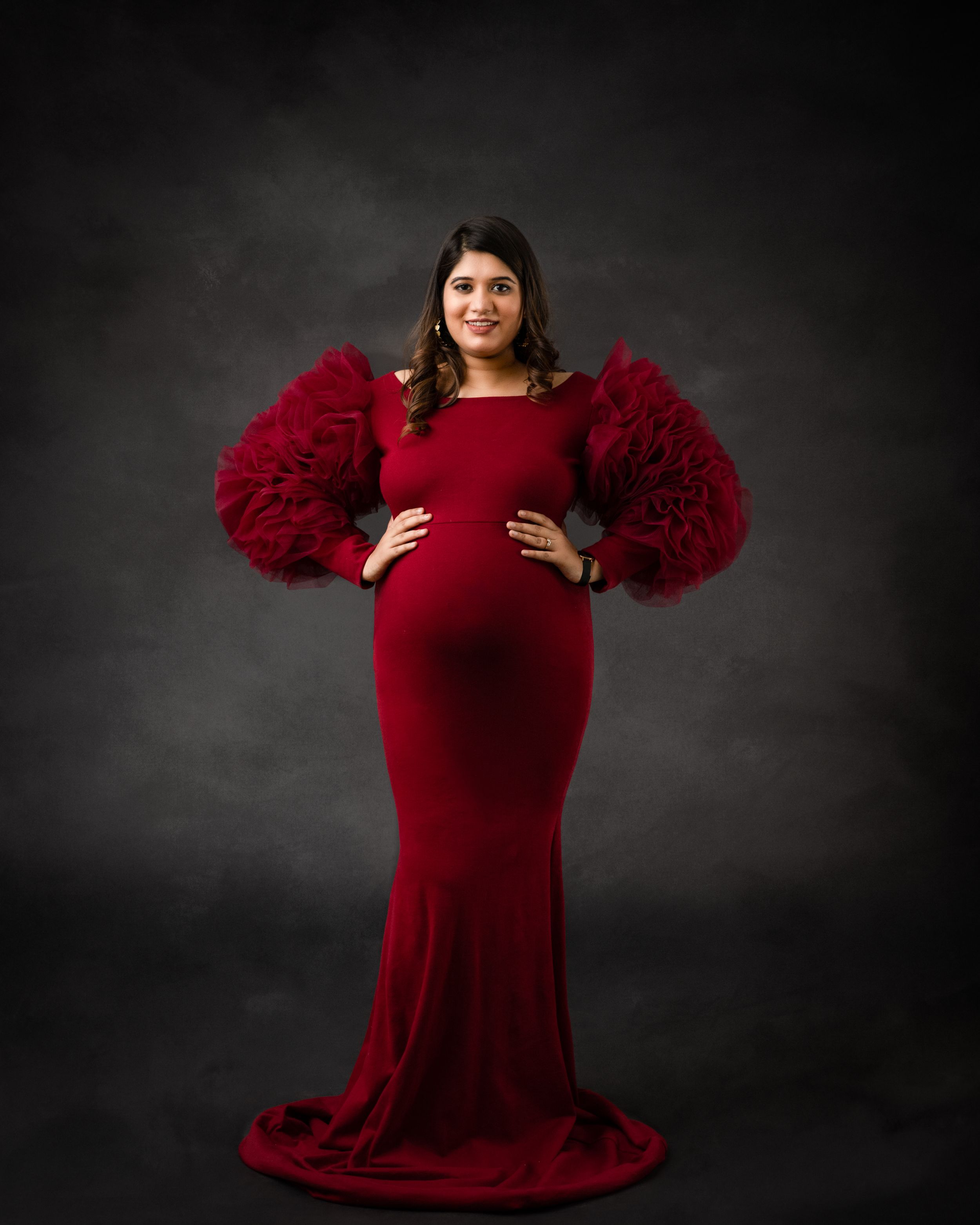 Dramatic maternity portrait in velvet red gown with puffy sleeves against dark background.