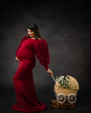 Maternity photo with person in red dress posing with decorative vintage baby carriage against dark backdrop.