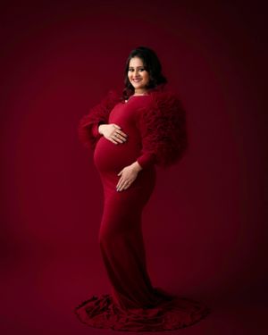Elegant maternity portrait featuring a floor-length red dress with dramatic ruffle sleeves against a deep red background.