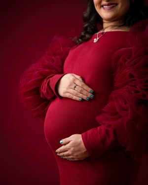 Maternity photo showing hands cradling a baby bump in a luxurious red dress with ruffled sleeves against burgundy backdrop.