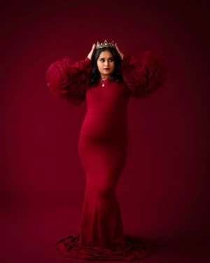 Maternity portrait in red dress with crown and dramatic sleeves against deep burgundy background.