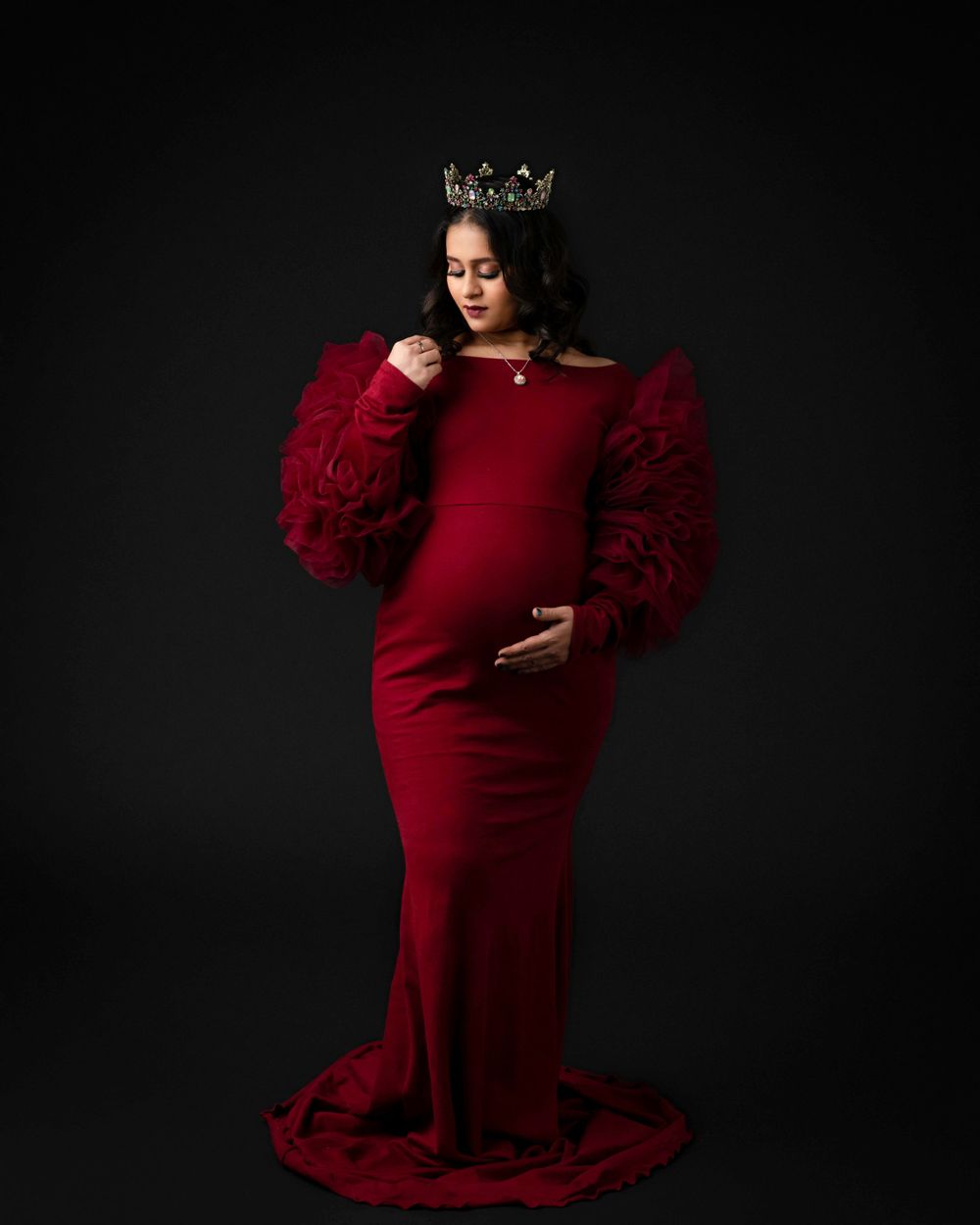 Elegant maternity portrait in deep red dress with puff sleeves against dark background.