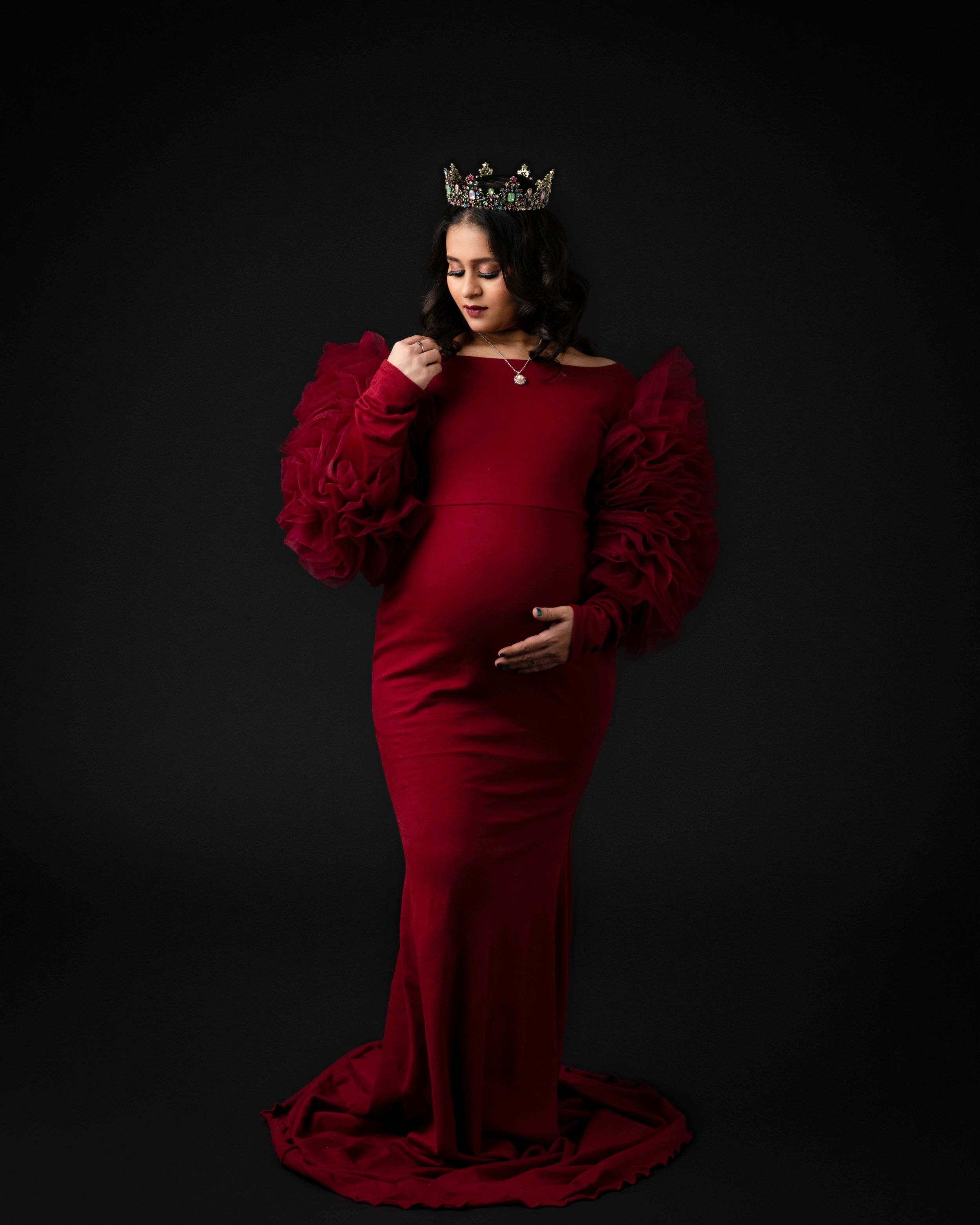 Elegant maternity portrait in deep red dress with puff sleeves against dark background.