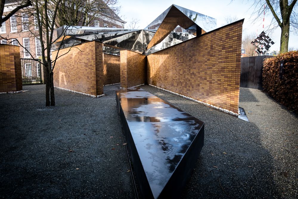 Modern memorial architecture featuring brick walls with angular mirrored steel roof panels outdoors.