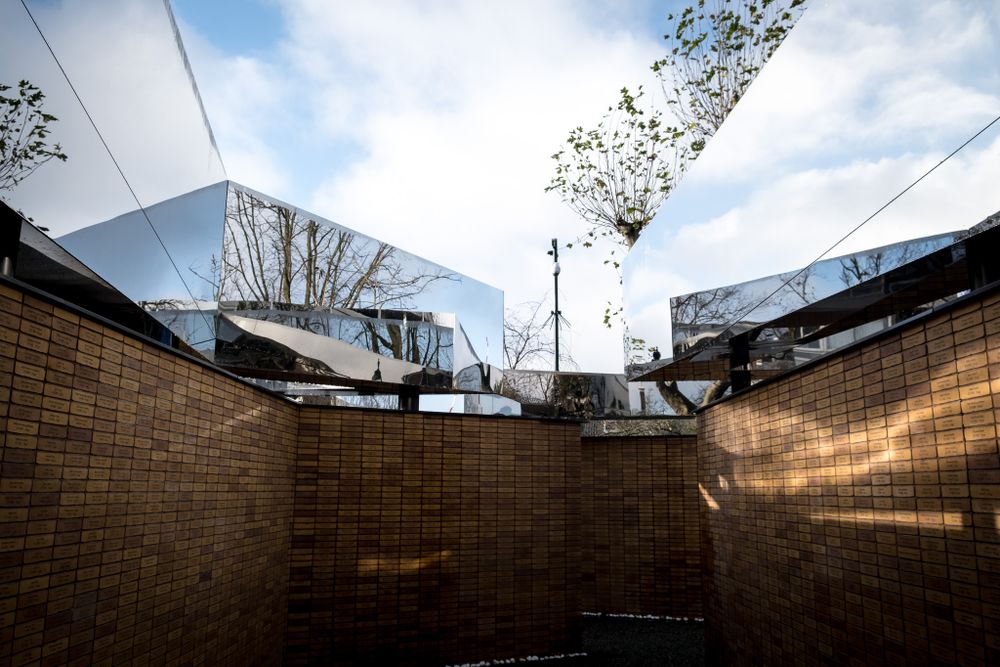 Mirrored geometric architecture with brick walls reflecting trees and sky in an urban courtyard.