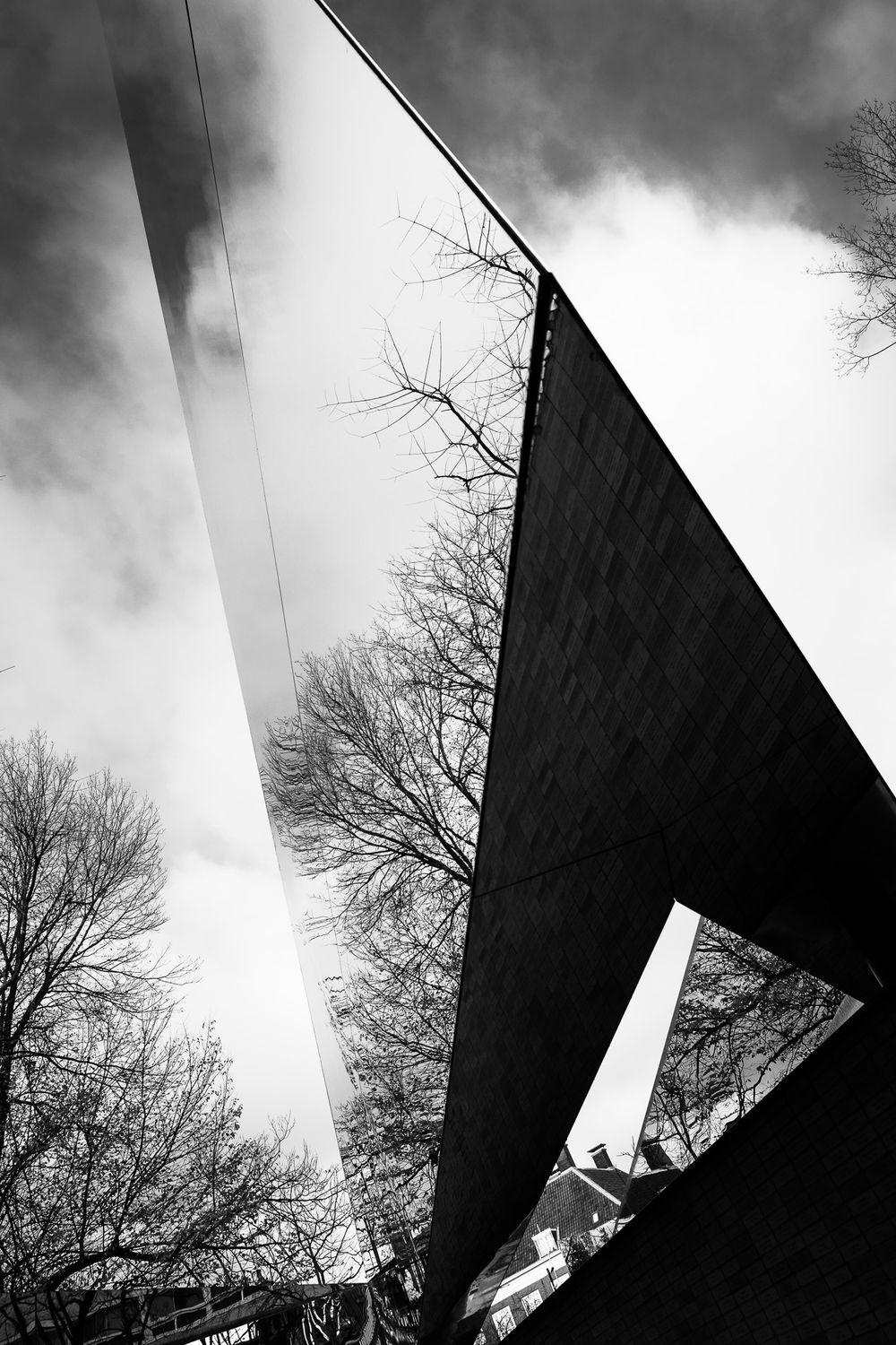 Low angle black and white view of a mirrored architectural structure with bare trees and dramatic sky.
