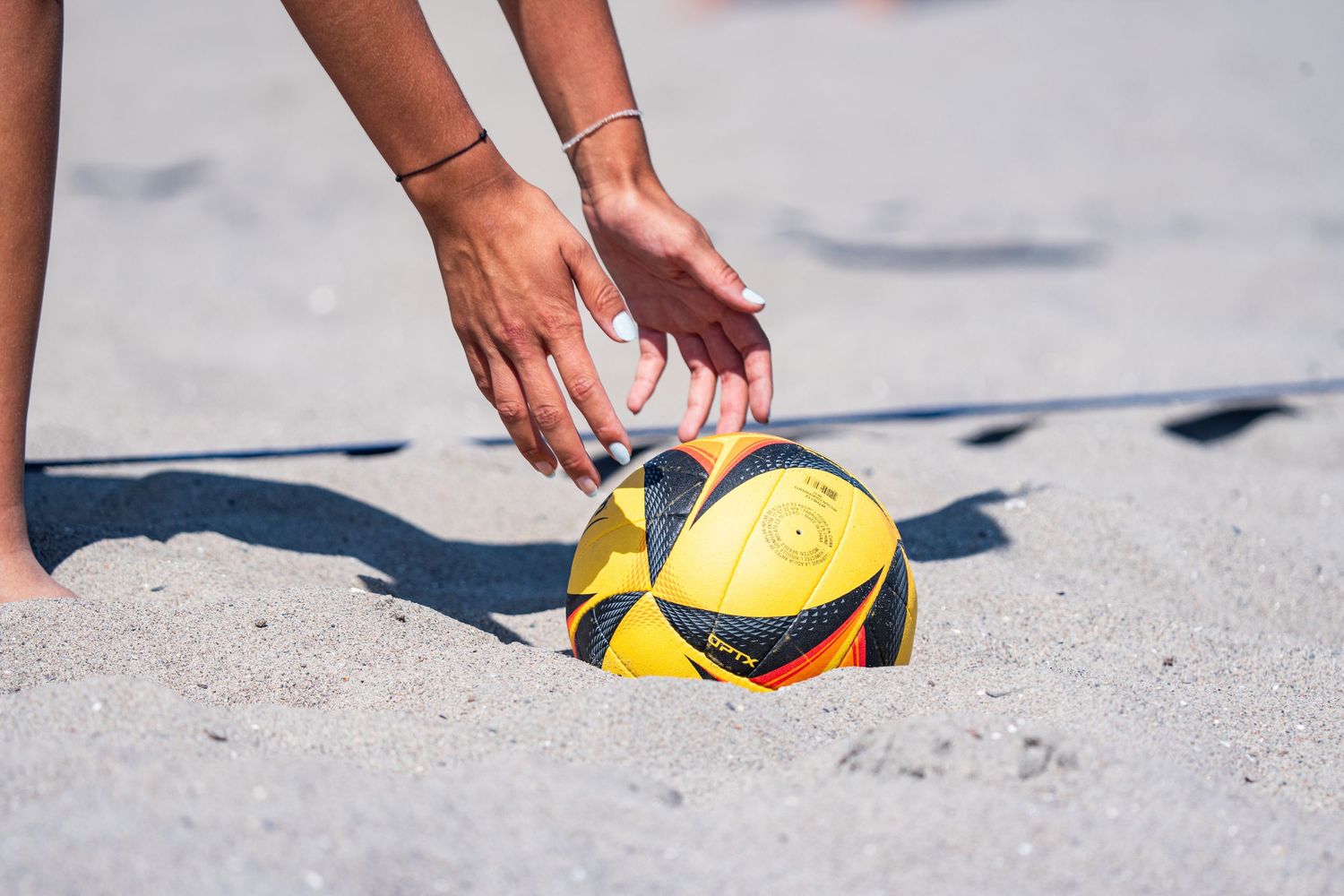 Hands reaching down to grab yellow volleyball on sandy beach surface.