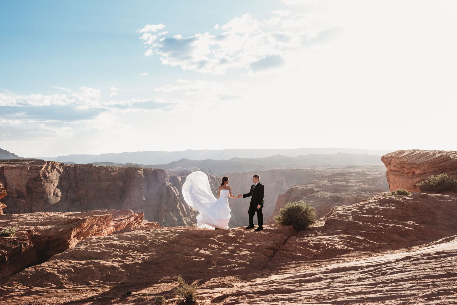 A couple stands together on red rock cliffs with dramatic canyon views.