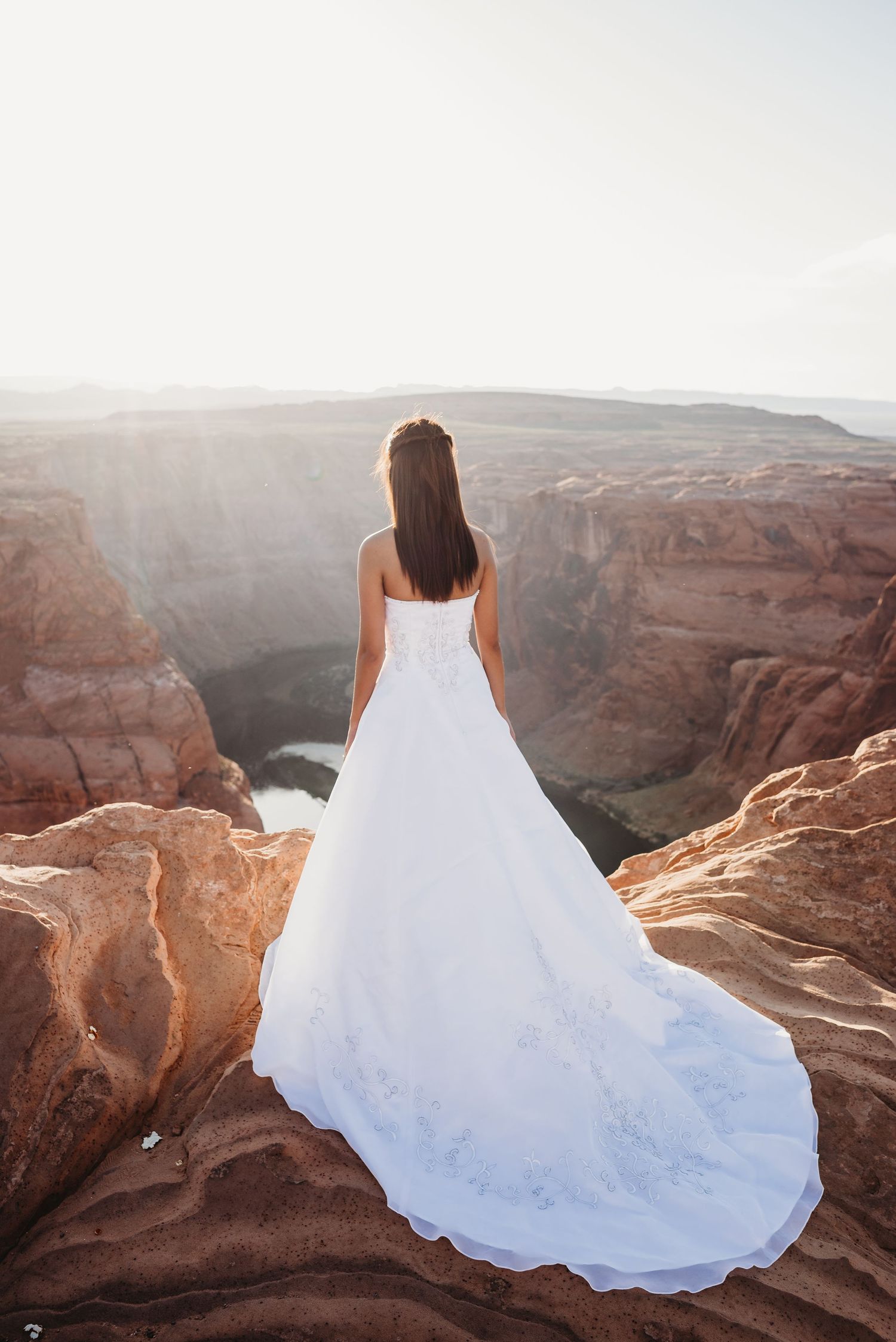 A white wedding dress flows dramatically against red canyon cliffs at sunset.