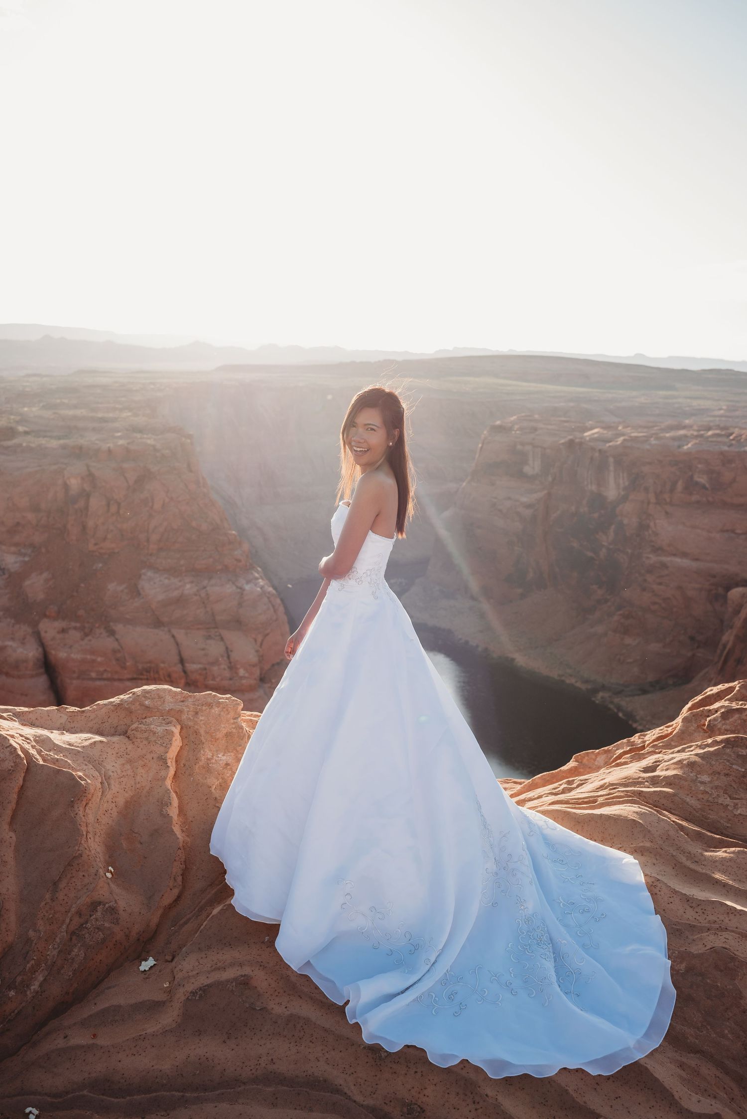 A flowing white wedding dress trails behind as a figure poses on red rock cliffs.