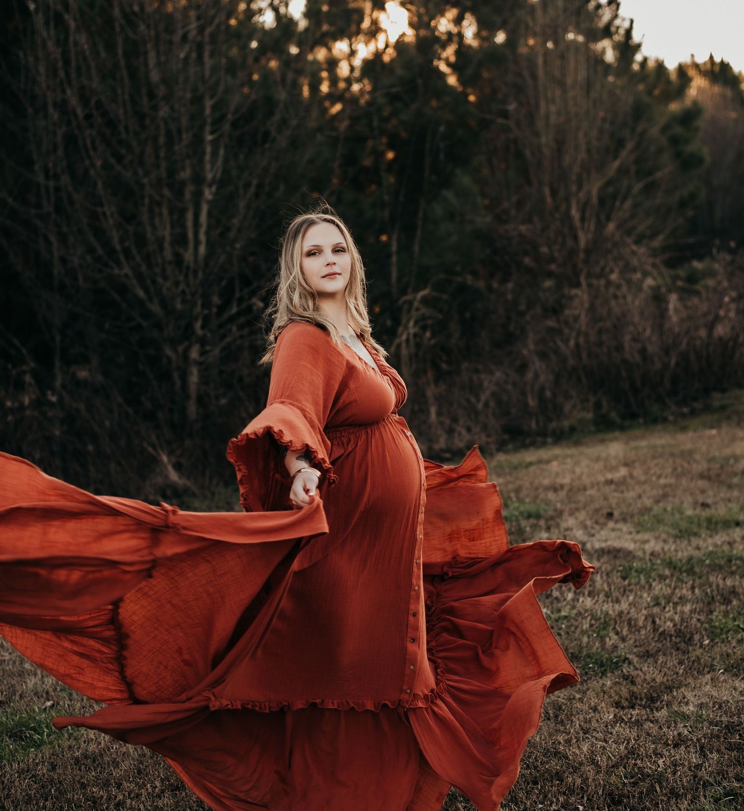 Pregnant woman in flowing rust-colored maxi dress poses dramatically in an outdoor autumn field.