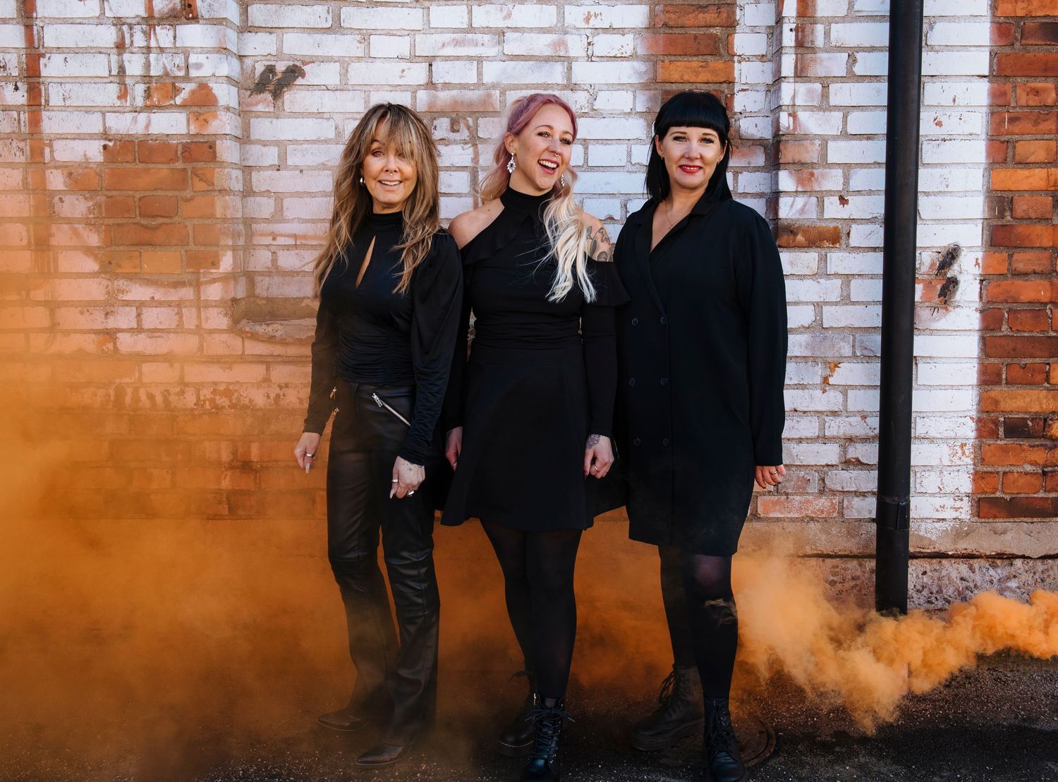 Three women in black outfits pose against a brick wall with orange smoke in the background.