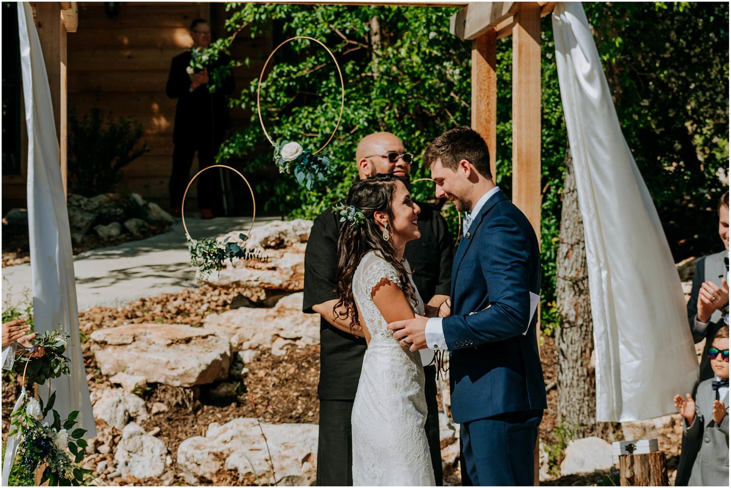 A joyful exchange during a wedding ceremony under a wooden pergola with white draping.