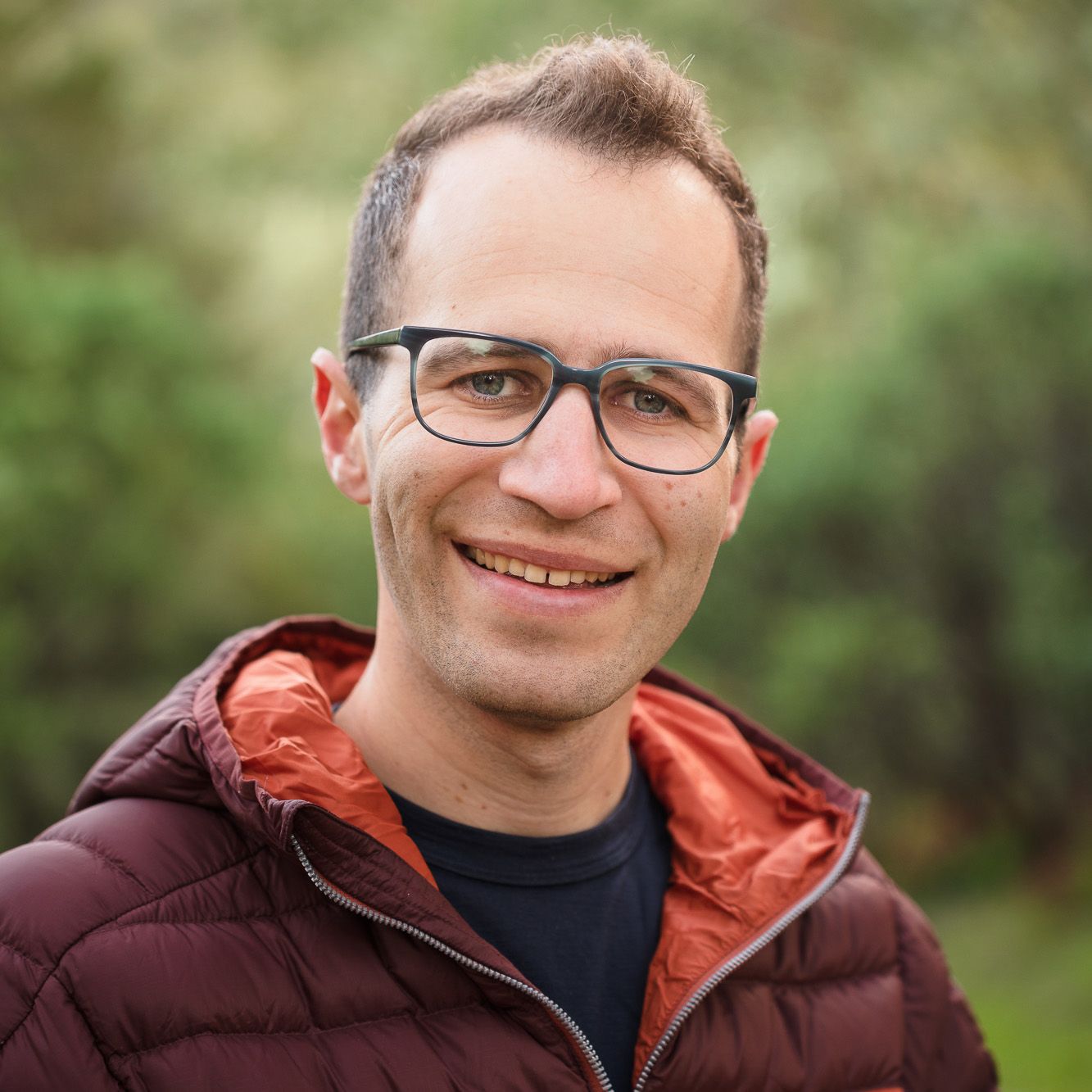Professional outdoor portrait of someone wearing glasses and a burgundy puffer jacket against a blurred natural background.