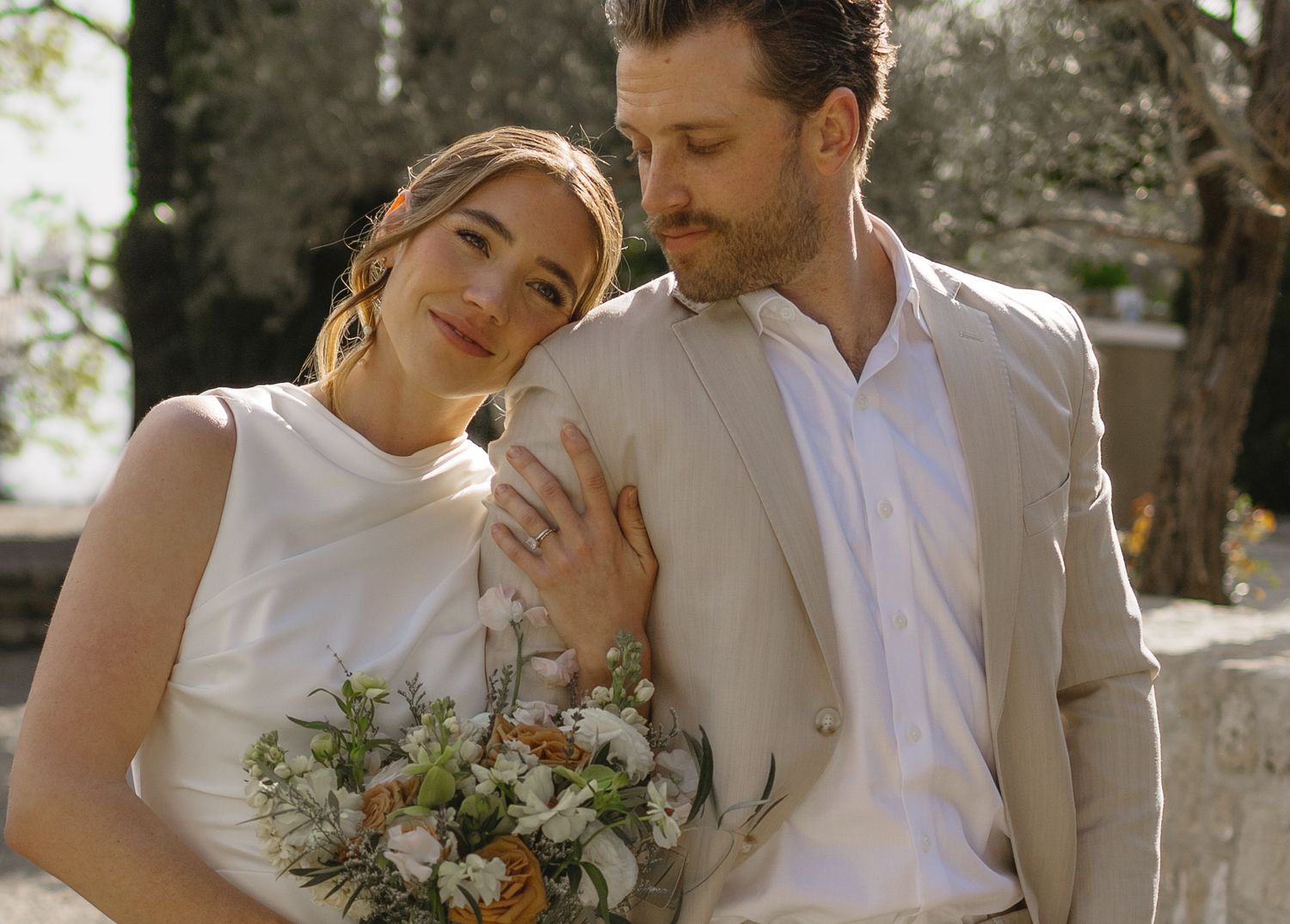 A romantic wedding couple shares an intimate moment outdoors with a beautiful floral bouquet.