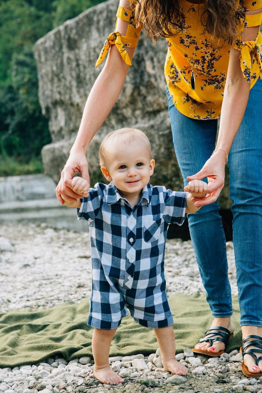 A toddler in plaid clothing holds hands while standing on a rocky shore.